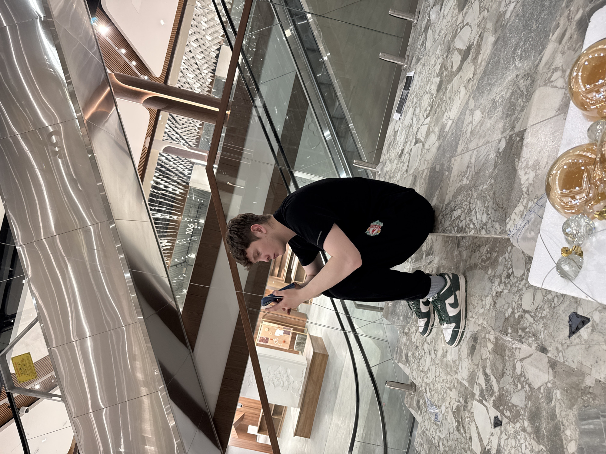 A young man is depicted in a modern, multi-level indoor environment, likely a shopping mall or large commercial building in Kazan, Russia. He is in a crouched or squatting position, focused on a dark-colored smartphone he holds with both hands. He appears to be in his late teens or early twenties, with short brown hair, wearing a black t-shirt adorned with the red Liverpool FC crest, black trousers, and green and white Nike Dunk sneakers with grey socks.
The setting features a striking architectural design with polished, reflective metallic surfaces, multiple glass railings forming transparent barriers, and warm-toned wooden or wood-paneled elements. White walls with decorative, possibly geometric, patterns are visible in the background, along with a number "106" on a vertical panel. The floor is made of large, irregularly shaped pieces of light grey and white marble or terrazzo.
On the right side of the image, close to the foreground, there are decorative items: several large, spherical, golden-hued glass ornaments and smaller, clear crystal-like decorations resting on a white surface. A small, yellow warning sign is faintly visible on a lower level in the background, behind the glass railings, though its text is not legible. The lighting is bright and artificial, characteristic of an indoor public space, creating reflections on the shiny surfaces. The man's posture suggests concentration on his phone.