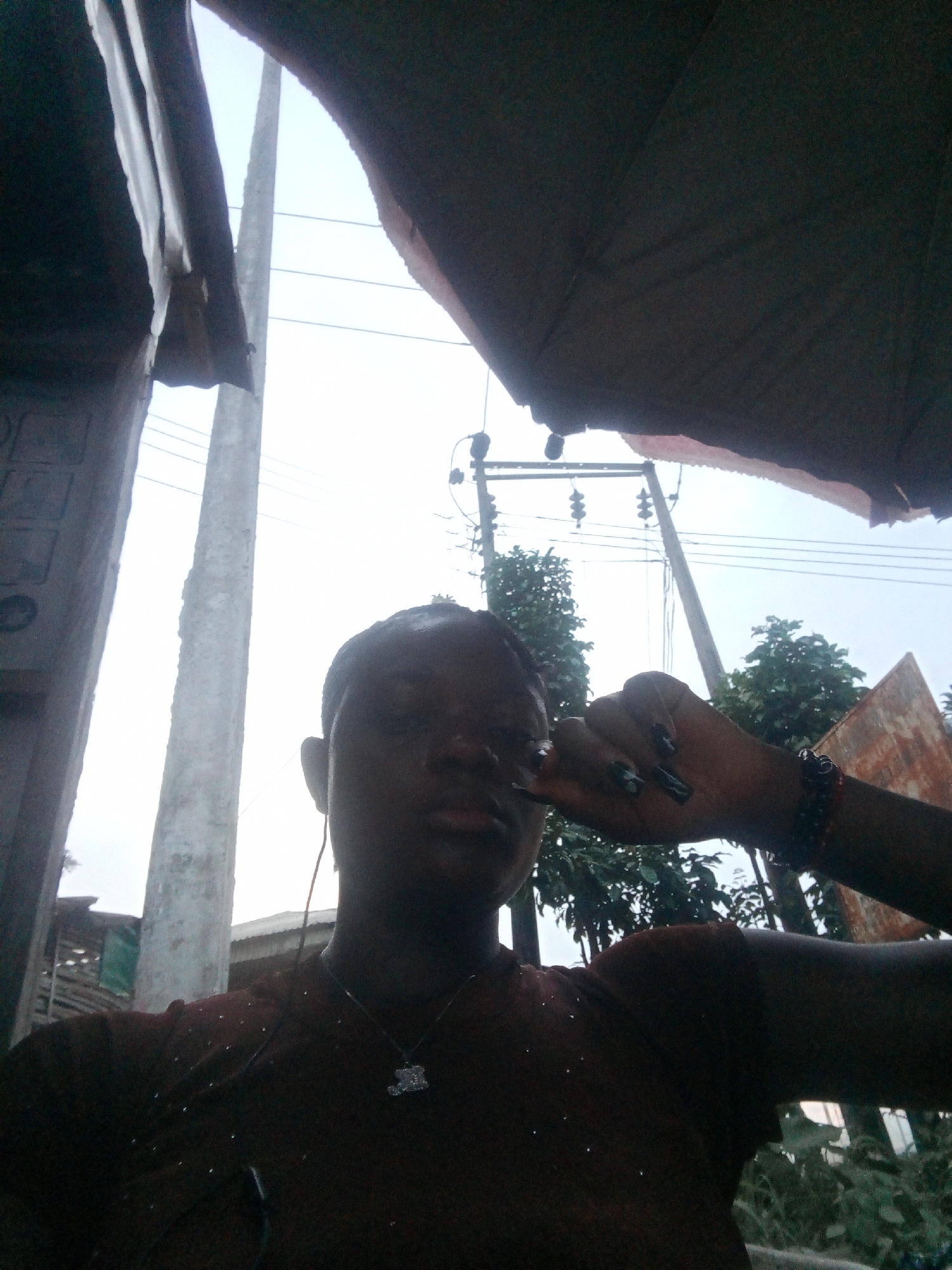 A low-angle, back-lit outdoor portrait captures a person, likely female, in Mbribit, Nigeria, during daylight hours. The subject is centered in the frame, looking towards the camera. Their face is largely in shadow against the bright, clear sky, obscuring facial features. They wear earphones, a dark-colored shirt with subtle light-colored specks, and a necklace with a small, light-colored charm, possibly a teddy bear. Their right hand, adorned with dark nail polish and a bracelet, is raised to the side of their head.
The background is dominated by a bright sky. Two large utility poles stand prominently, one on the left and another slightly right of center, supporting multiple power lines that stretch across the upper mid-ground. Lush green trees and foliage are visible beneath the power lines. Further in the background, elements of structures, including what appears to be a corrugated metal roof, can be vaguely discerned. The foreground is framed by dark, ambiguous overhead structures, possibly an awning or temporary shelter, creating a natural vignette and contributing to the strong backlighting on the subject. The scene depicts a casual moment in a public, semi-urban or residential outdoor setting. No discernible text is visible.