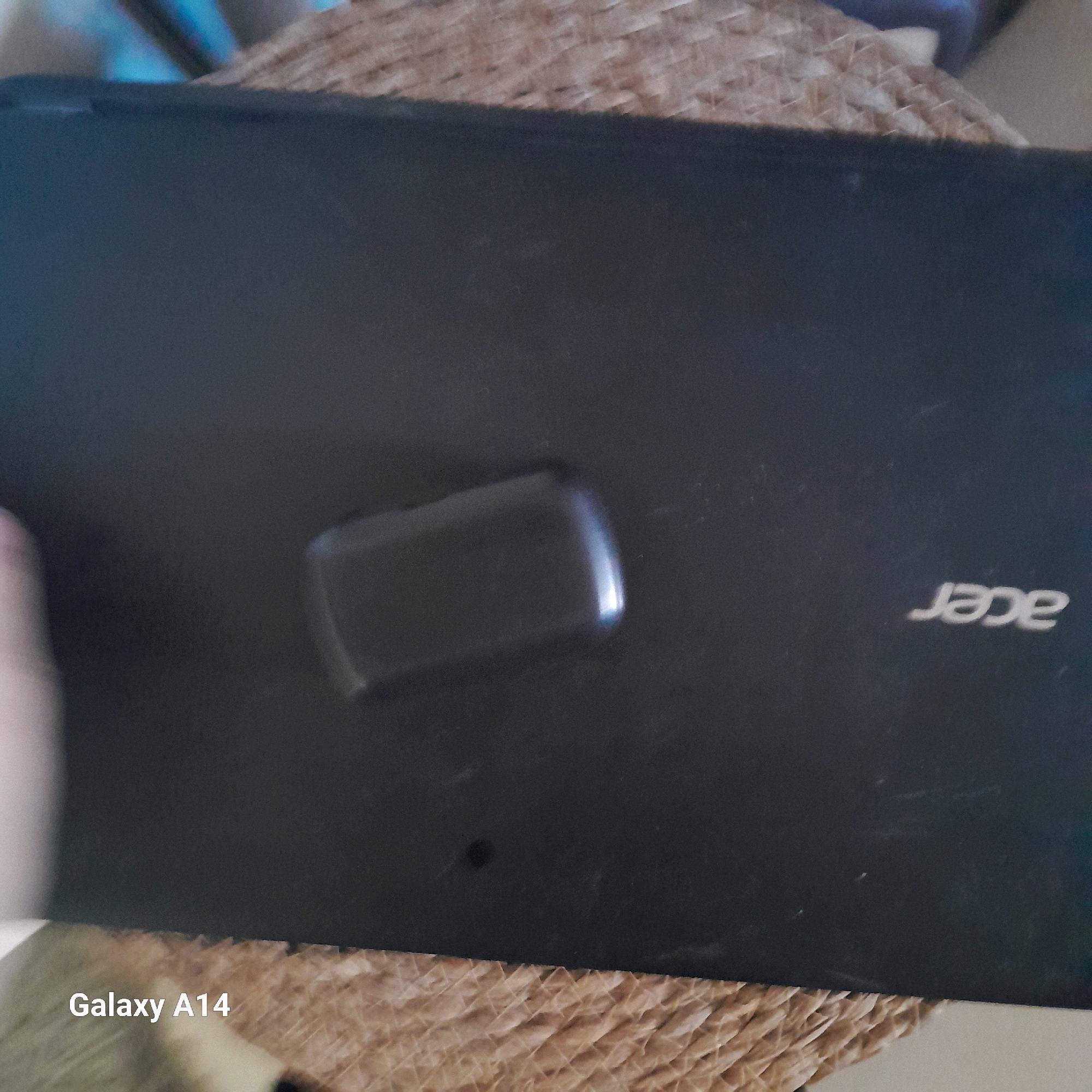 A dark, slightly reflective laptop lid is positioned on a woven, textured surface. The laptop lid has a slightly recessed, rectangular area in the center, which is also reflective. The brand logo "acer" is visible in white text on the right side of the laptop lid. In the bottom left corner of the image, the text "Galaxy A14" is superimposed in white. The overall scene appears to be an indoor setting, but the specific location within Khan Yunis, Palestinian Territories, cannot be determined from the visual information provided in the image. There are no people or explicit actions depicted.