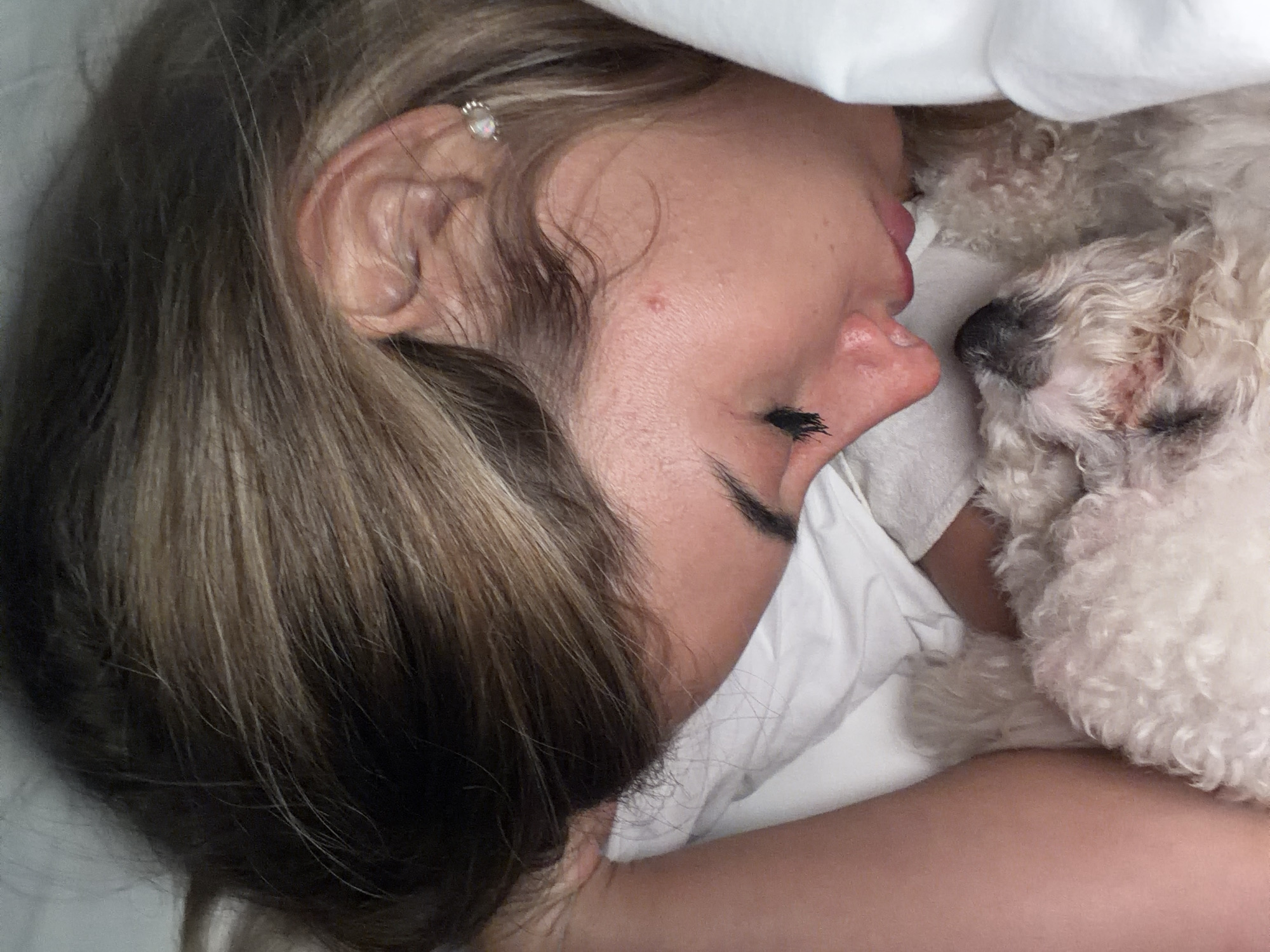 A woman lies in bed, her head resting on a white pillow next to a small white dog. She is sleeping, with her eyes closed and her dark eyelashes visible. Her hair is highlighted and parted to the side. She has an earring in her ear. The dog is also sleeping, nestled close to the woman's face. The scene takes place in an indoor setting, presumably a bedroom. The location is not discernible from the image provided.