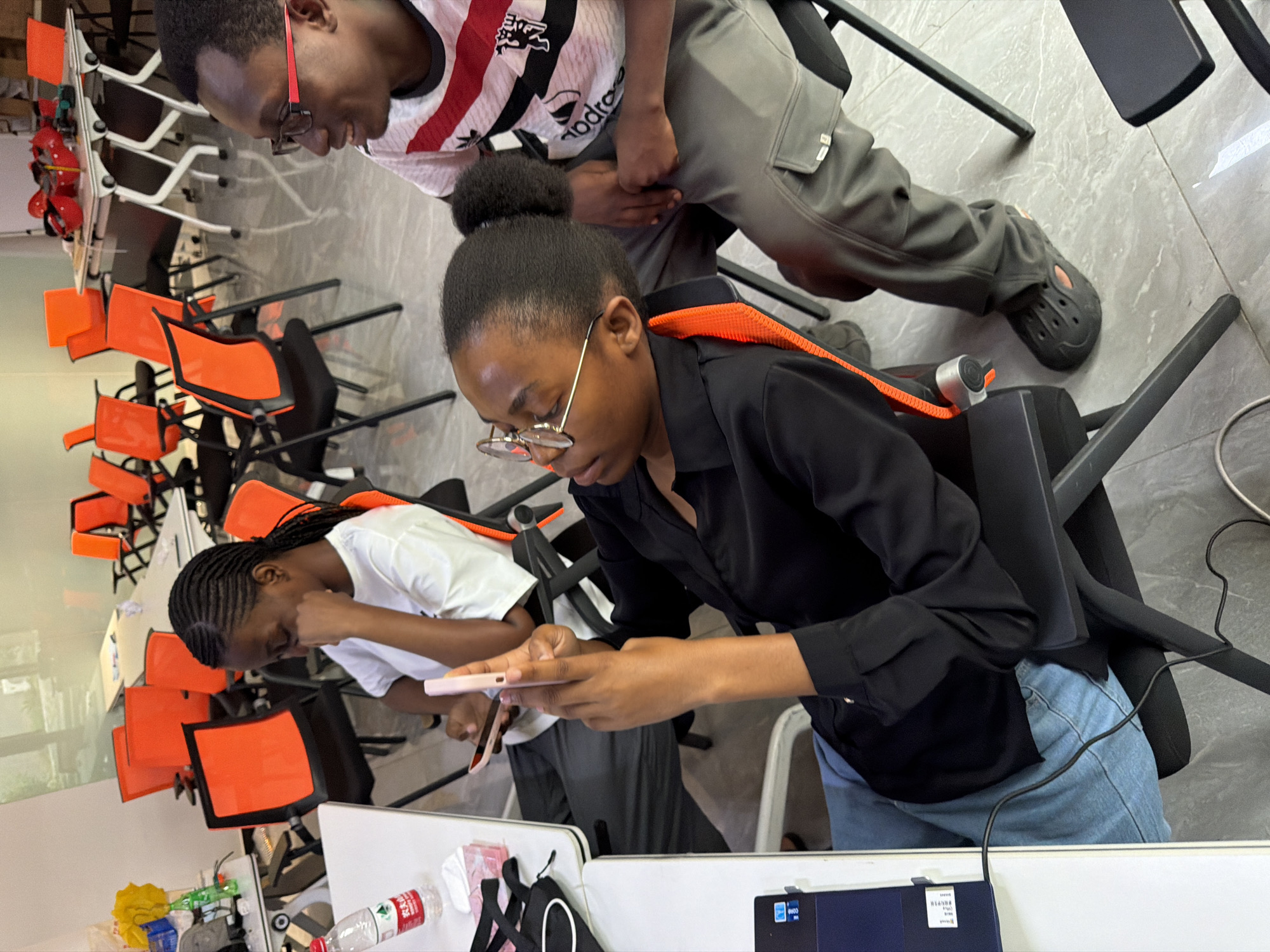 In an indoor setting resembling a classroom or meeting room, three young Black individuals are seated at desks. The room is equipped with multiple orange-backed chairs and white tables. In the foreground, a young woman with glasses and dark hair pulled back is holding a pink smartphone. She is wearing a black collared shirt and blue jeans. To her left, another young woman with braided hair is also looking at a smartphone, wearing a white t-shirt. To the right, a young man wearing glasses and a red and white striped t-shirt is looking away from the camera. A laptop is visible on a desk in the foreground. The environment appears to be clean and organized, with a tiled floor. The overall scene suggests a casual and informal gathering or study session.