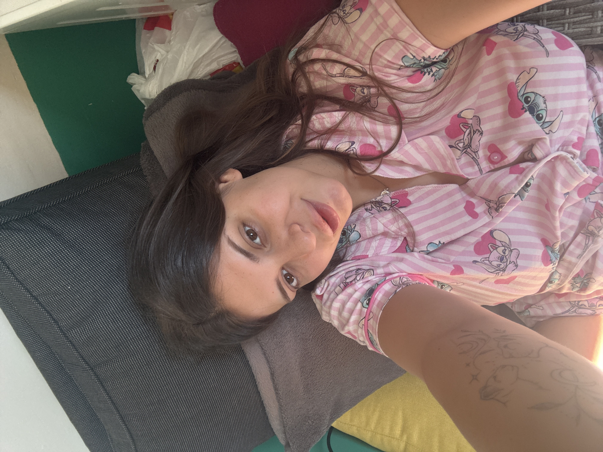 A young woman with long, dark hair and brown eyes is lying on her side, looking at the camera. She is wearing pink and white striped pajamas with Stitch characters and pink hearts. Her left arm, visible in the foreground, has a tattoo. She is positioned on a couch with several pillows, including a dark grey one, a lighter grey one, and a yellow one. Behind her, a green wall is visible, along with some white plastic bags and a red object. The lighting suggests it is daytime, possibly indoors. The image does not contain any discernible elements that would allow for the localization to Plovdiv, Bulgaria.