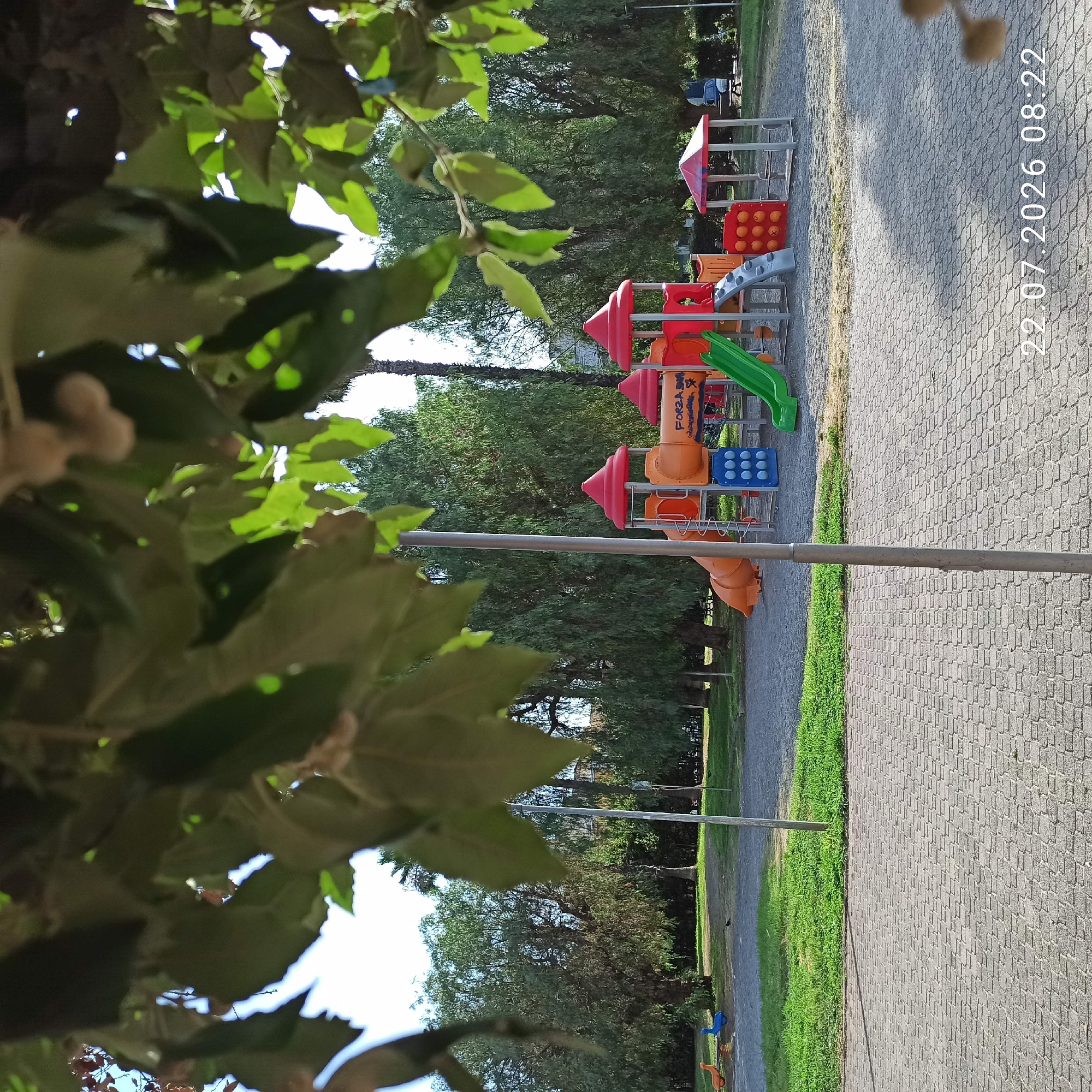 The image depicts a children's playground located in the city of Modugno, Italy, as indicated by the date stamp "22.07.2026 08:22". The playground features a multi-colored play structure with slides, tunnels, and climbing elements. Orange and red are prominent colors in the play structure, which also has blue circular panels and grey steps. In the background, a line of trees forms a natural backdrop, with some palm trees visible among them. The playground area is bordered by a paved walkway made of interlocking grey bricks. A patch of green grass separates the paved area from the playground's soft ground covering. To the left of the main play structure, there are two smaller, brightly colored children's rocking toys, one orange and one blue. A tall, thin metal pole stands in the foreground, slightly off-center. The sky is clear and bright, suggesting daytime. There are no people actively playing in the playground at the time the photo was taken.