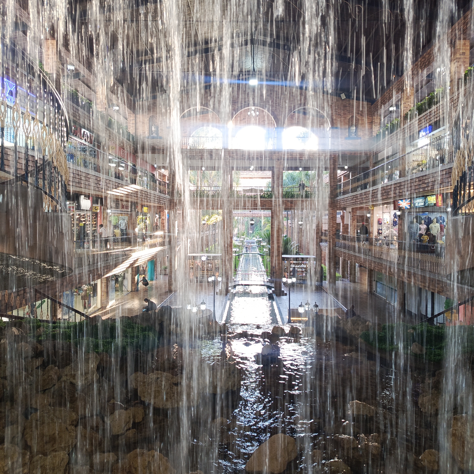 The image shows a view through a curtain of falling water into an indoor courtyard or atrium of a multi-story building. The architecture features exposed brick, arched windows, and multiple levels of walkways and shops. Sunlight streams in from the far end of the atrium, illuminating a long, narrow water channel with bridges crossing it. People are visible on the walkways, in the shops, and near the water features. The foreground is dominated by rocks and the flowing water. This location appears to be the Sambil Paraguan in Carrizal, Venezuela.