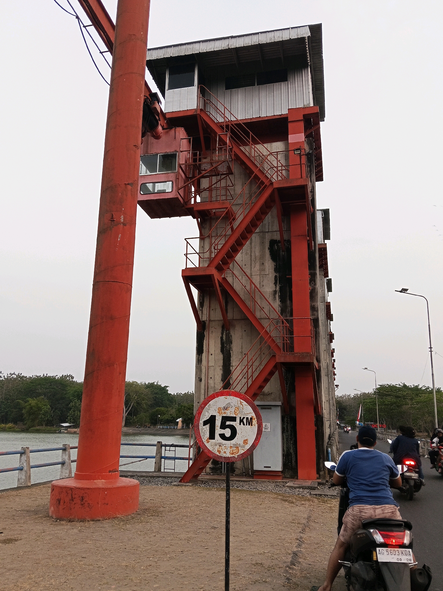The image depicts a tall, red and gray industrial structure, possibly a water management facility, located in Gogodeso, Indonesia. The structure features multiple levels, external metal staircases painted red, and large windows. In the foreground, a person on a scooter wearing a blue shirt and beige shorts is visible from behind, facing away from the camera and riding on a paved road. A speed limit sign with "15 KM" is prominently displayed in front of the structure. Beyond the structure, a body of water, trees, and distant buildings can be seen under an overcast sky. Other people on motorcycles are traveling along the road in the background, suggesting a public area. The overall scene conveys a sense of an operational industrial site within a community setting.