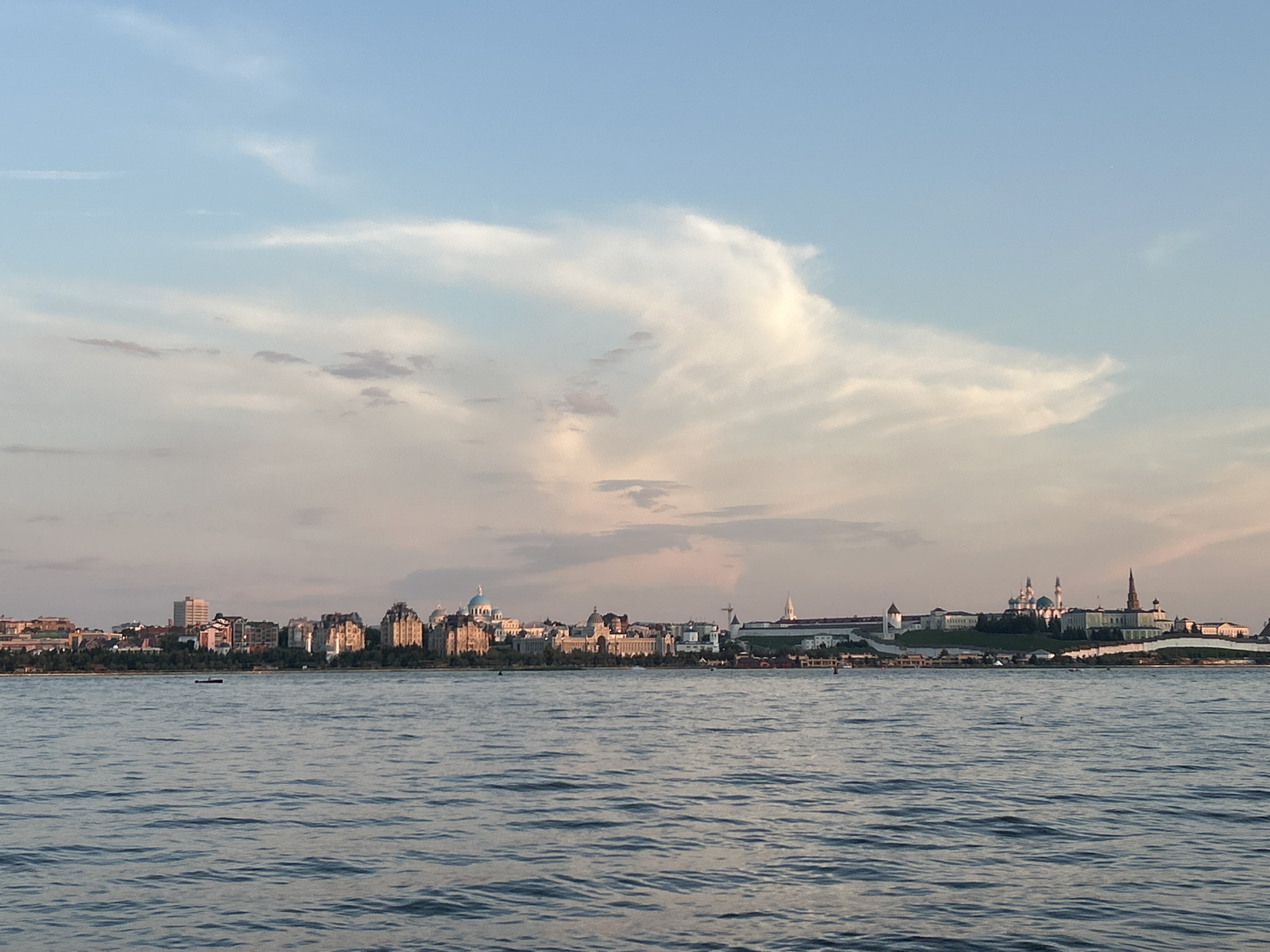 The image captures a serene view of the Kazan Kremlin across the Volga River. In the foreground, the wide, calm river reflects the soft hues of the sky. The water has a rippled texture, suggesting a gentle breeze. A small, dark, rectangular object, possibly a buoy or a small boat, is visible on the water in the mid-ground.
The middle ground features a long, low shoreline lined with trees. Beyond the trees, the iconic silhouette of the Kazan Kremlin rises against the horizon. Prominent structures include several domes, including a distinct blue-domed cathedral, and minarets, indicative of the complex's rich religious and historical significance. The Kremlin buildings appear to be a mix of white stone and darker materials, with various architectural styles visible. White walls and fortifications are also noticeable, encircling the complex.
The sky above is a gradient of soft blues and pinks, transitioning from a clear blue at the top to warmer tones near the horizon, suggesting it is either sunrise or sunset. Wispy, ethereal clouds are scattered across the sky, adding depth and texture to the scene. The overall lighting is soft and diffused, casting a gentle glow on the city and the water. No people are clearly identifiable in the image. The scene evokes a sense of tranquility and historical grandeur.