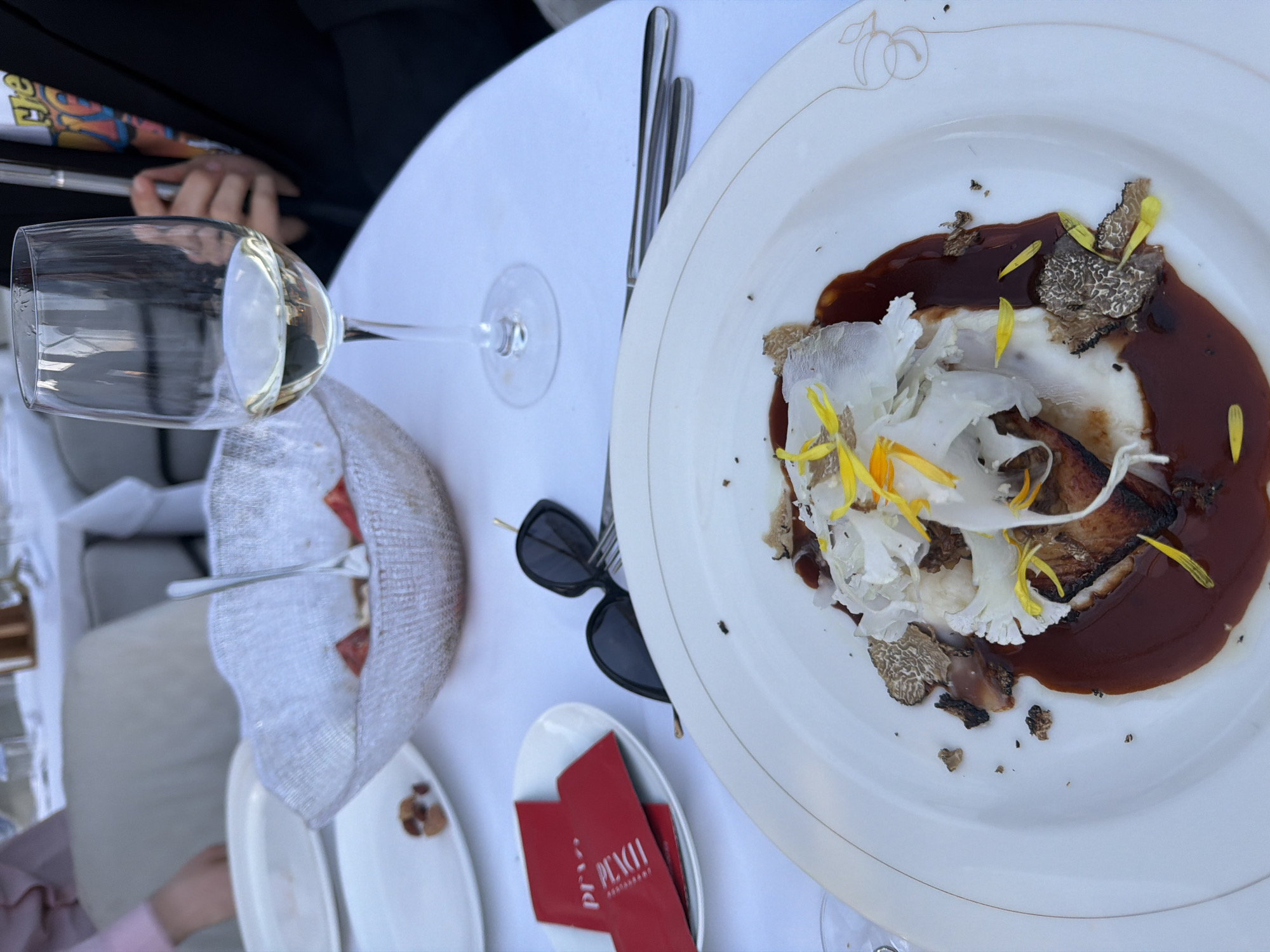 This media file presents an overhead view of an elegantly set table at what appears to be an outdoor restaurant, identified as "PI PEACH RESTAURANT" located in Moscow, Russia. The scene is brightly lit by natural daylight, suggesting a pleasant daytime meal.
The central subject is a gourmet dish served on a white oval plate. The dish features a seared piece of food, possibly meat or fish, on a creamy white base, complemented by delicate, thin white shavings (which could be vegetables like cauliflower or certain mushrooms), rich dark brown sauce, and generous slices of black truffle. It is artfully garnished with vibrant yellow flower petals. The plate itself bears a subtle golden logo on its rim, depicting two peaches, aligning with the restaurant's name.
To the left of the main dish, a wine glass holds a light-colored liquid, likely white wine. A person's hand, partially visible with a dark sleeve in the upper left, is near the glass. Below the wine glass, a white fabric-lined bread basket contains reddish items, possibly fruit or bread. Another hand, wearing a pink sleeve, is visible towards the bottom left.
Other objects on the white table include a pair of black sunglasses, a fork and knife, and two small white oval dishes. One of these dishes holds two red cards, clearly displaying the text "PI PEACH RESTAURANT" in white, confirming the establishment. The activity depicted is people dining and enjoying a refined meal in a relaxed, al fresco setting.