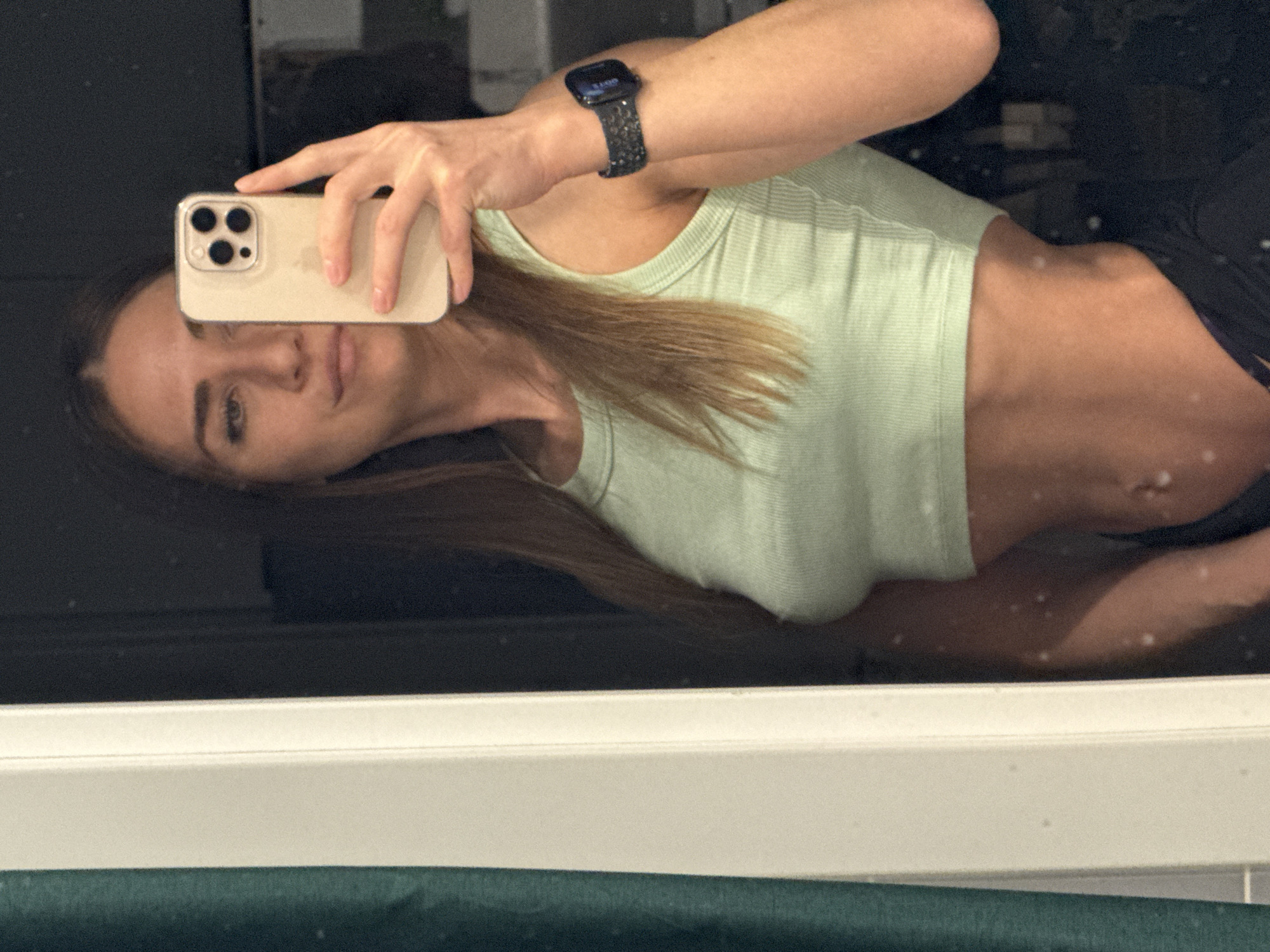 A woman is taking a selfie in a mirror. She's wearing a light green ribbed tank top and a smartwatch on her left wrist. Her phone is a modern iPhone with multiple rear cameras. The environment appears to be a bathroom with dark wall paneling and a white countertop, possibly a shared or public restroom. The location in Lyubertsy, Russia, cannot be confirmed from the visual information provided.
