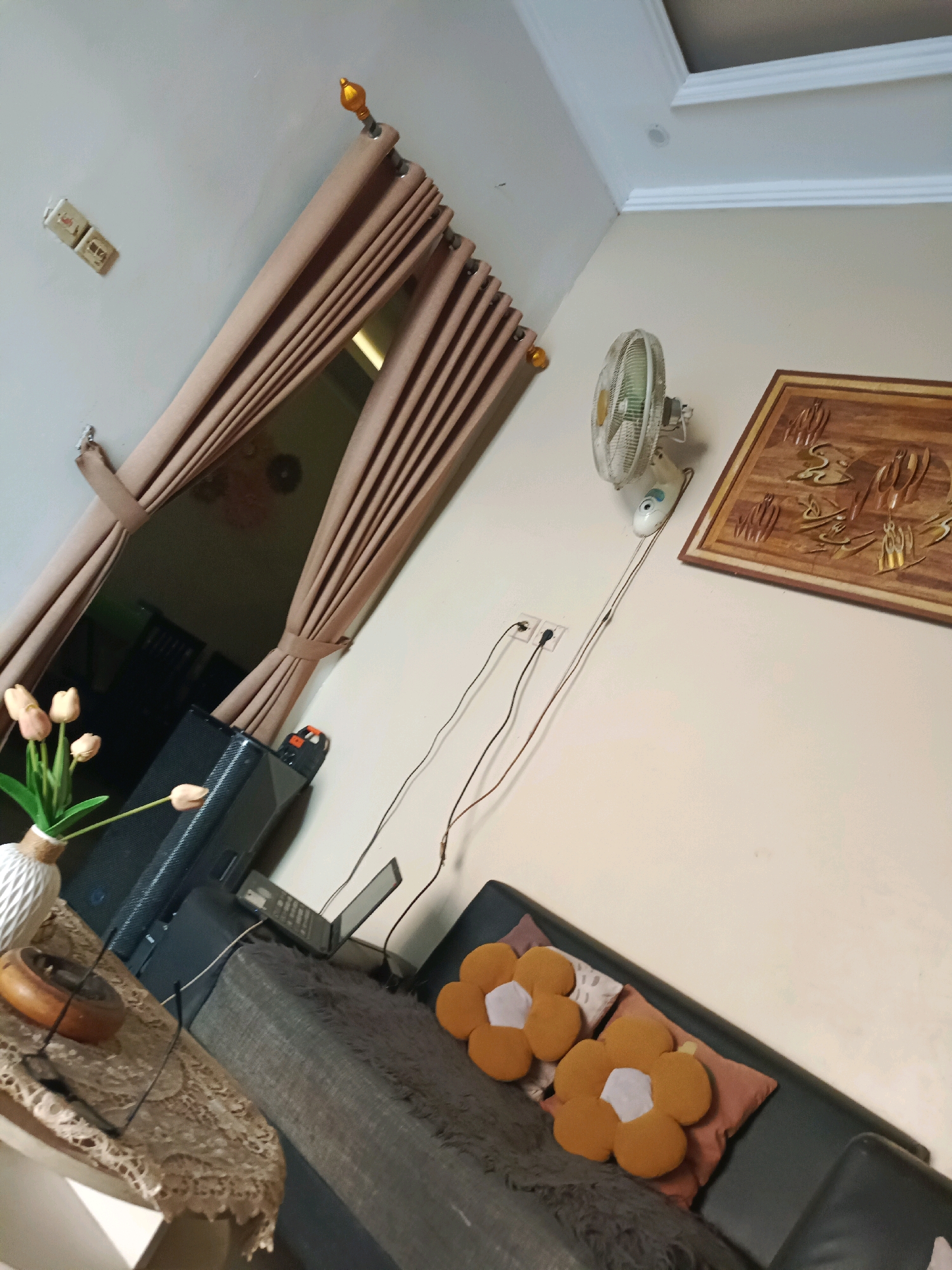 The image depicts an indoor scene, likely a living room, in the city of Rancang Kencono, Indonesia. A brown curtain is drawn back to reveal a dark interior space. On the wall, a white oscillating fan is mounted above a framed wooden artwork with intricate carvings. To the left of the fan, electrical outlets and cords are visible. In the foreground, a black sofa is partially visible, adorned with two orange flower-shaped cushions and a dark gray throw blanket. A small laptop is placed on the sofa. To the left, a vase with artificial tulips sits on a table covered with a lace tablecloth, next to a dark speaker. The walls are painted a light neutral color, and a white ceiling with decorative molding is present at the top of the frame.