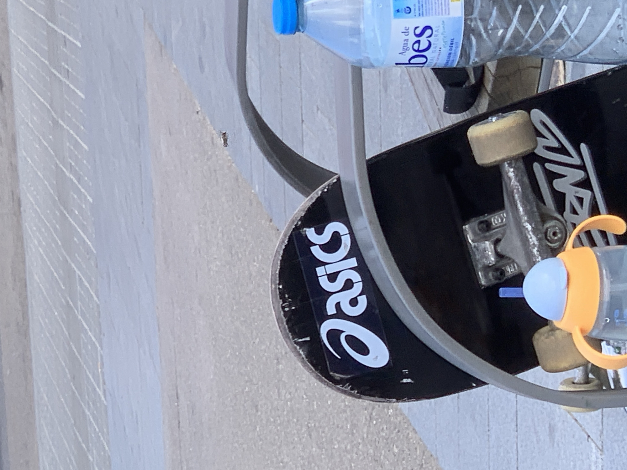 The rotated image presents a close-up of a skateboard and two bottles resting in an outdoor paved area during the daytime. No people are visible in the frame.
The black skateboard deck features a prominent white "Asics" sticker on its tail or nose. Part of another white graphic or logo is also visible on the main body of the deck. The skateboard is equipped with light-colored wheels and silver trucks, showing some signs of wear and use. It appears to be leaning against or placed on a curved metal rail or structure.
Positioned near the skateboard are two drinking containers. A clear plastic water bottle with a blue screw cap is visible, featuring a label that clearly reads "Agua de CABES" and "NATURAL", indicating it is natural water. Below it, an infant's sippy cup is present, characterized by its bright orange handles, blue spout, and clear body.
The ground surface is a light gray or beige pavement, possibly concrete or large tiles, exhibiting a subtle patterned texture. The bright, even lighting suggests a sunny day. The scene depicts a casual outdoor setting in el Prat de Llobregat, Spain.