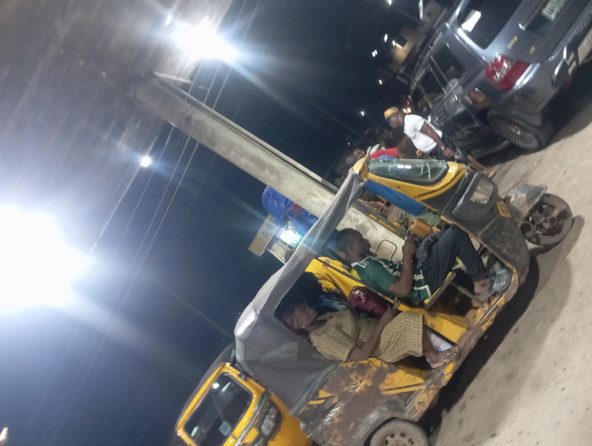 The media file captures a night scene in Nkpor, Nigeria, featuring a yellow and black three-wheeled auto-rickshaw, commonly known as a Keke Napep, and a grey SUV. The image is taken from an angled perspective, with prominent lens flares from bright artificial lights illuminating the dark sky and creating a glow on a large concrete pillar or overhead structure that traverses the upper left.
Inside the Keke Napep, a man in a green and white striped shirt is seated, while a woman in a light-colored, patterned traditional outfit appears to be resting. The vehicle itself shows noticeable wear and tear, with chipped paint and dirt. Behind the Keke, a grey SUV is parked, and a man in a white t-shirt and yellow cap stands near its rear. Other indistinct vehicles are visible in the background. The ground beneath the vehicles is a mix of paved and dusty surfaces. The scene suggests a busy urban environment, possibly a street or a taxi stand, late at night, with people either traveling or waiting. There is no clearly readable text on any of the vehicles or surrounding elements; a very faint sign with partially visible letters like "DIE" or "DIS" can be barely discerned in the background in some cropped views, possibly indicating a shop or a fuel station.