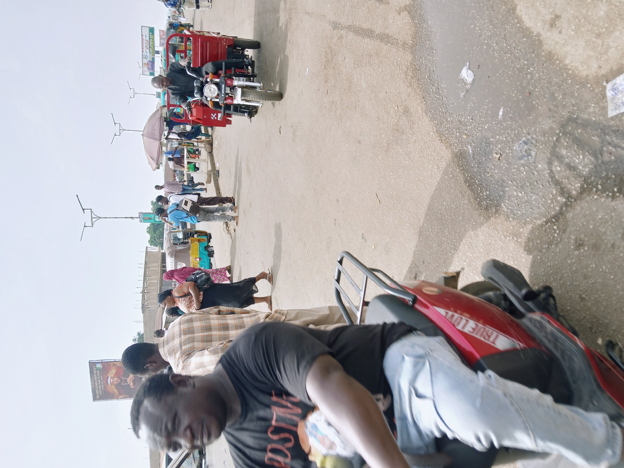 The image captures a street scene in Jos, Nigeria. A man in a black t-shirt and light blue jeans is seen in the foreground, seemingly near a red motorcycle. Further down the street, a group of people is visible, some standing near what appears to be market stalls or shops under a pale pink umbrella. A red tricycle with a driver is moving in the same direction. The street appears to be paved, with a visible puddle. A billboard featuring the words "WE ARE GENERATION NEXT AND THE TIME IS NOW" and "LOVE 2027" is situated on the side. Text visible on the red tricycle reads "DAYLONG".