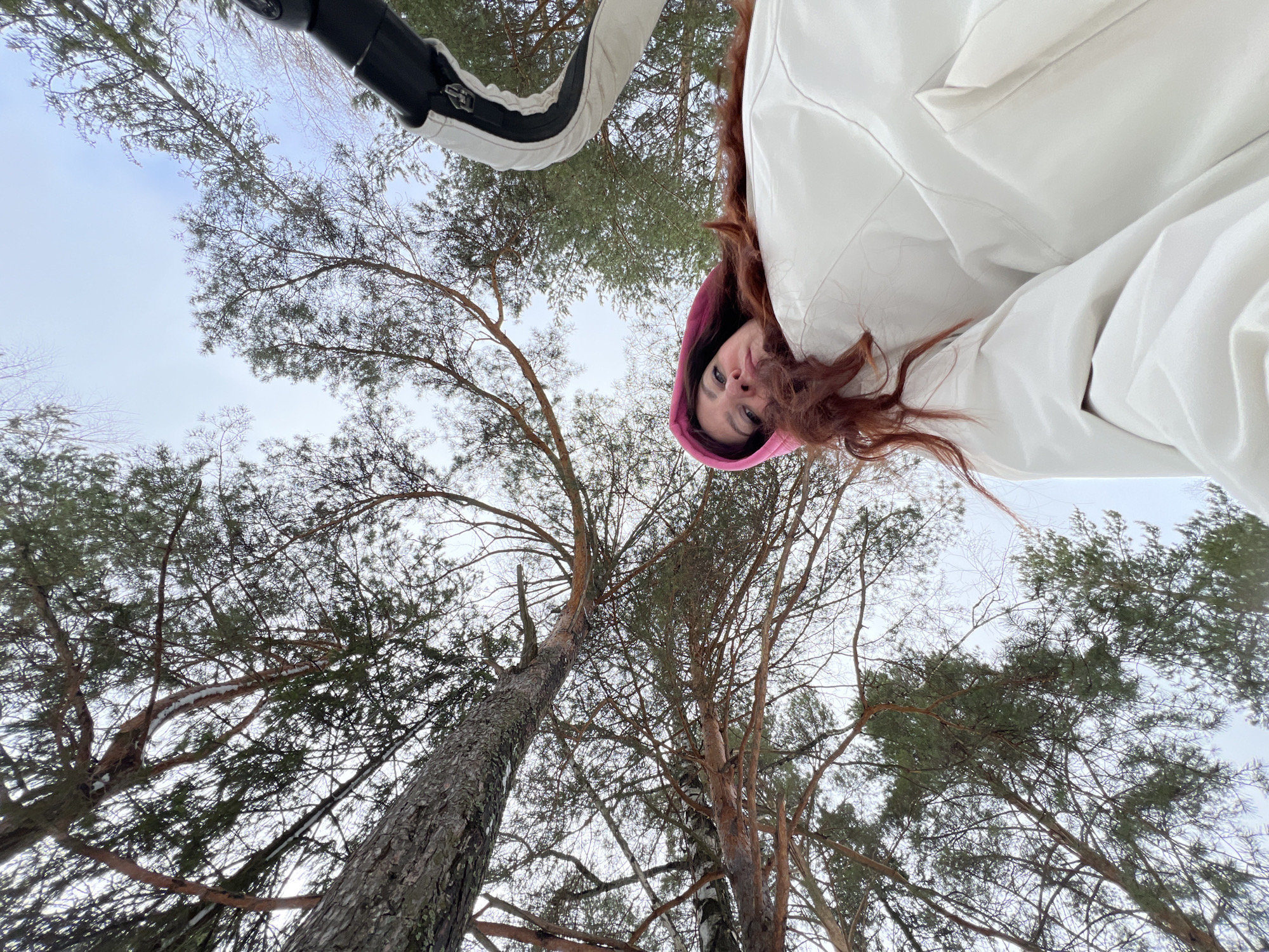 The image is a low-angle shot taken outdoors, likely in a forest or park in Noviy, Russia. A person is holding the camera, wearing a white jacket with a pink hood, partially visible in the upper right. Long reddish-brown hair frames their face. Above and behind the person, there are tall trees with bare branches and some with green foliage, silhouetted against a pale sky. Some branches have small patches of snow. In the upper left corner, a section of a white and black vehicle (possibly a snowmobile or ATV) is visible.
