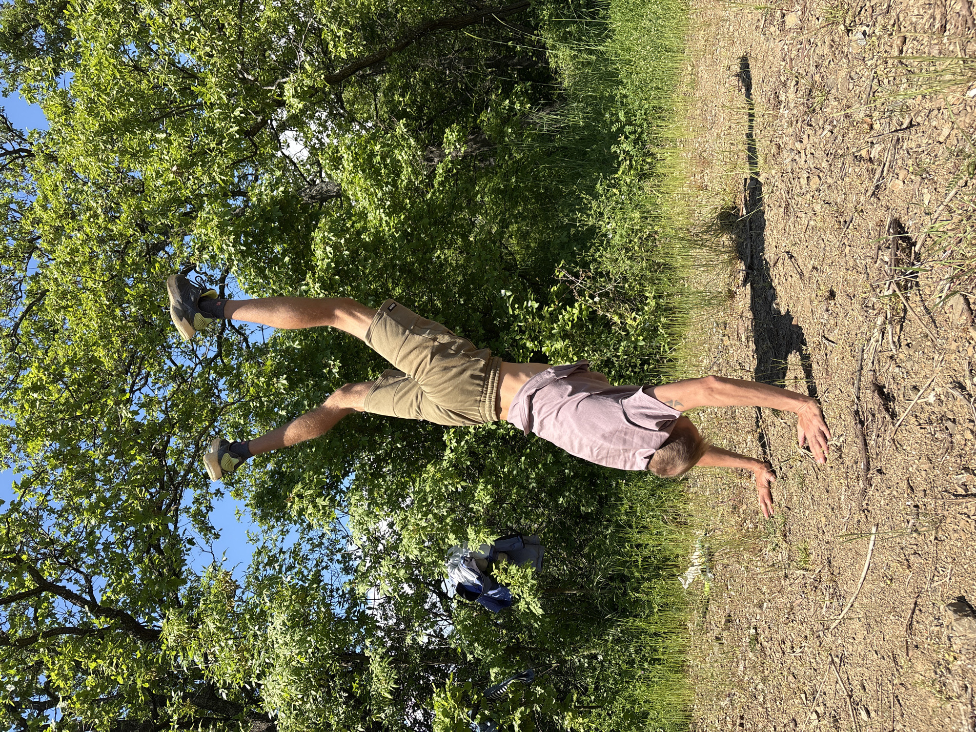 A man is performing a handstand in a natural environment. His body is oriented horizontally in the image frame, with his hands planted on a patch of ground and his legs extended upwards amidst foliage. He is wearing a light purple t-shirt and khaki shorts, with athletic shoes on his feet. The ground where his hands are planted appears to be a mix of dirt, small rocks, and sparse vegetation. Behind the man and his legs, there is a dense background of trees and green vegetation, suggesting a wooded or park area. A glimpse of blue sky is visible through the trees. A backpack and articles of clothing are visible between the trees. It is not possible to determine if the photograph was taken in the city of Nizhnee Zaprudnoe, Ukraine with the information provided.