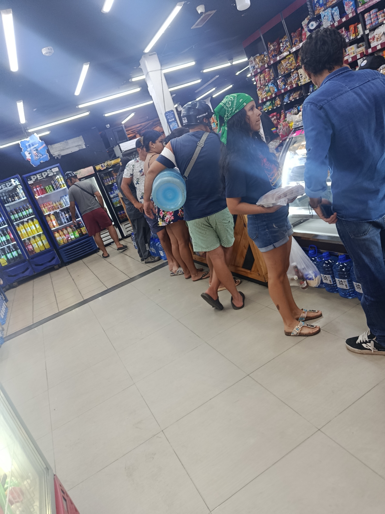 The image depicts a scene inside a store in Altagracia, Venezuela. A line of people is waiting, presumably to make a purchase. The people in the line are wearing casual clothes. A woman in a green bandana stands near the front of the line. Many of the customers are carrying large bottles of drinking water, likely for refilling. The store has shelves stocked with groceries and refrigerated cases filled with beverages. The lighting is bright from fluorescent ceiling lights.