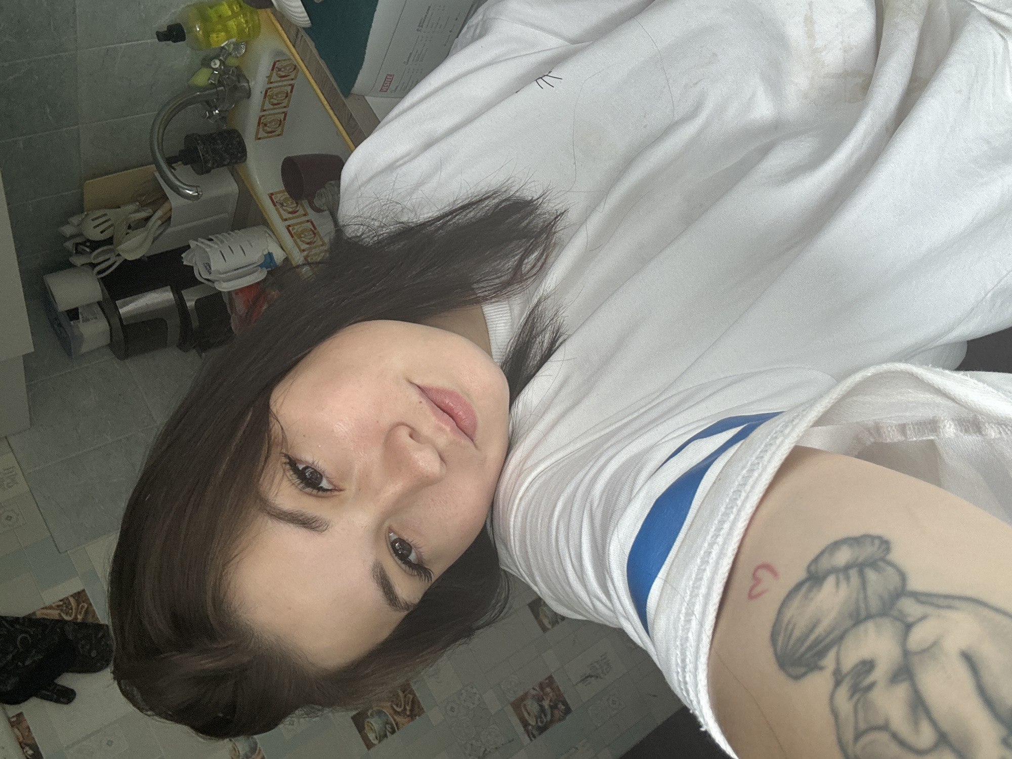 The image shows a young woman with brown hair and fair skin taking a selfie in what appears to be a kitchen. She is wearing a white t-shirt with blue stripes on the sleeve and has a tattoo on her arm. The background shows a kitchen sink with dishes, a faucet, and various kitchen utensils. The walls are tiled with a pattern that suggests they are in a residential setting. The lighting suggests it is daytime. The location is identified as Chaikovsky, Russia.