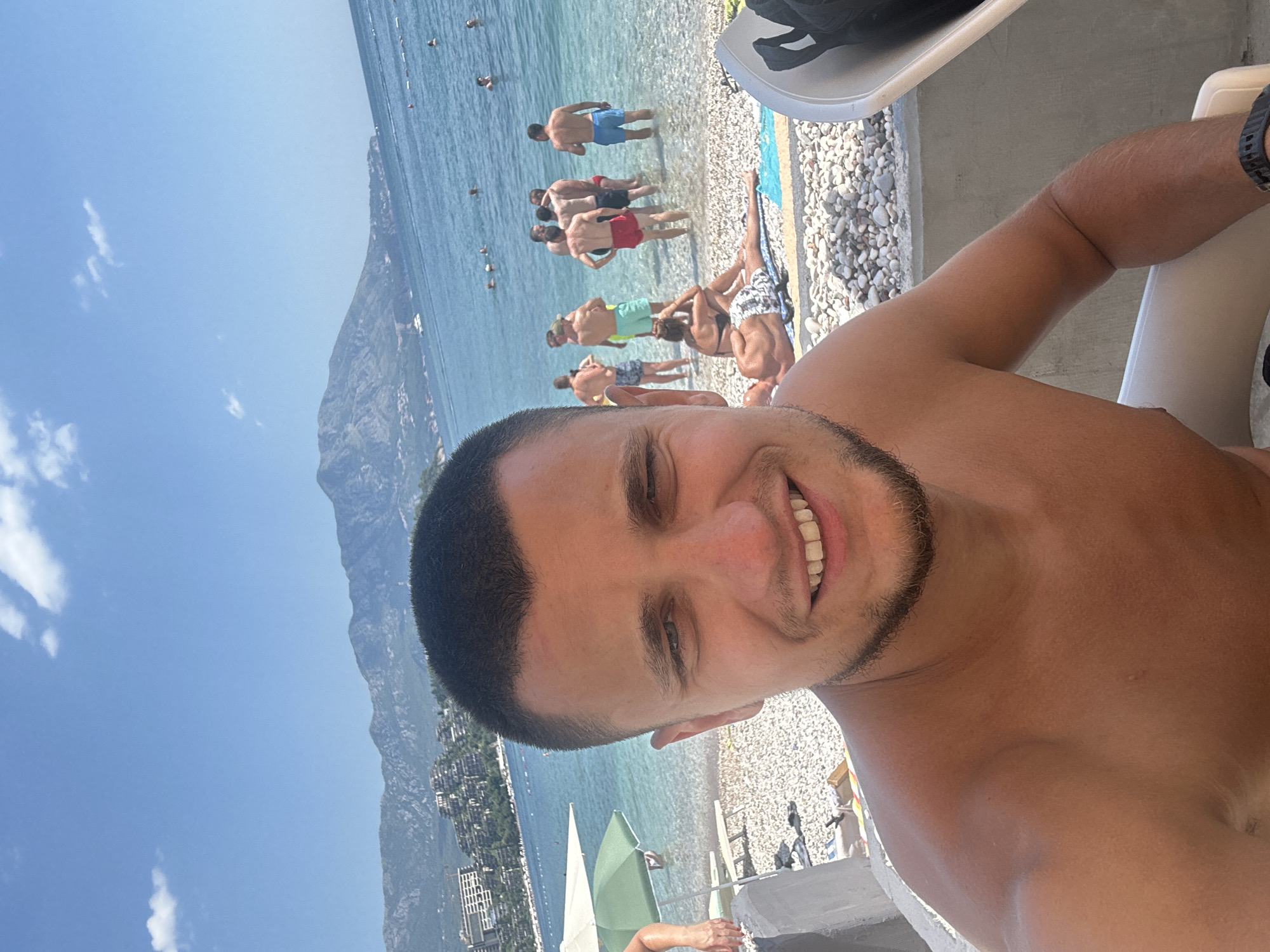 A man with short dark hair and stubble smiles at the camera, taking a selfie on a pebbly beach in Budva, Montenegro. He is shirtless, wearing a dark watch on his left wrist. In the background, the clear blue Adriatic Sea is dotted with swimmers. Behind the sea, a mountainous coastline is visible, with buildings clustered on the slopes. Several green and white beach umbrellas are scattered across the shore, and lounge chairs are present. A group of men are standing in the shallow water, some in swim trunks. Another person is lying on a towel on the beach. The sky is bright blue with a few scattered clouds.