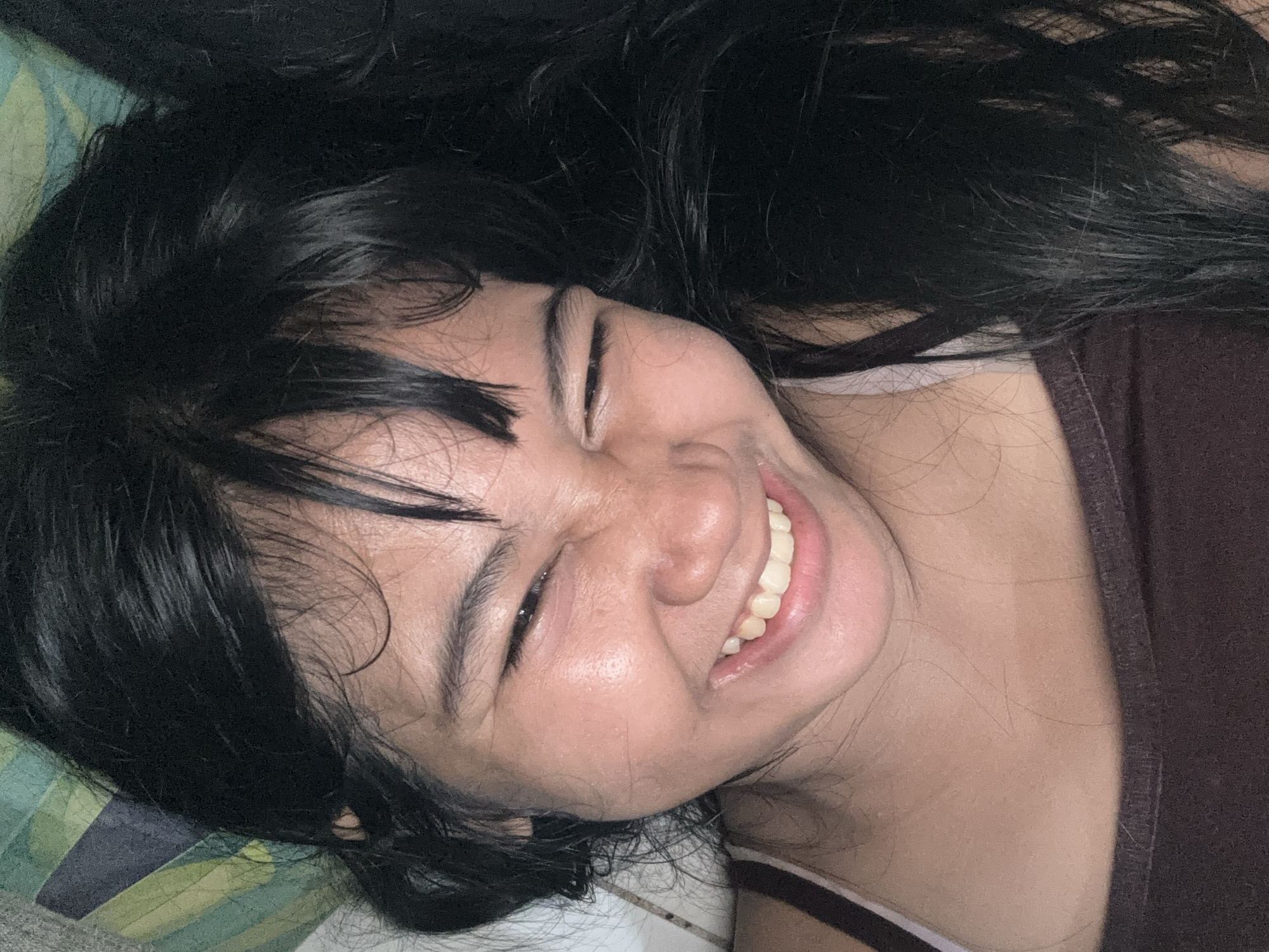 A close-up, head-and-shoulders shot captures a young woman with dark hair laughing, her eyes squinted shut and her mouth wide open, revealing her teeth. She is wearing a dark brown tank top with a white strap visible. The background is a soft focus of green and blue, possibly a pillow or blanket. The lighting suggests an indoor setting, perhaps a bedroom, and the angle of the shot indicates she is lying down. The image appears to be a candid moment, taken from a low angle. The photograph is from North Jakarta, Indonesia.