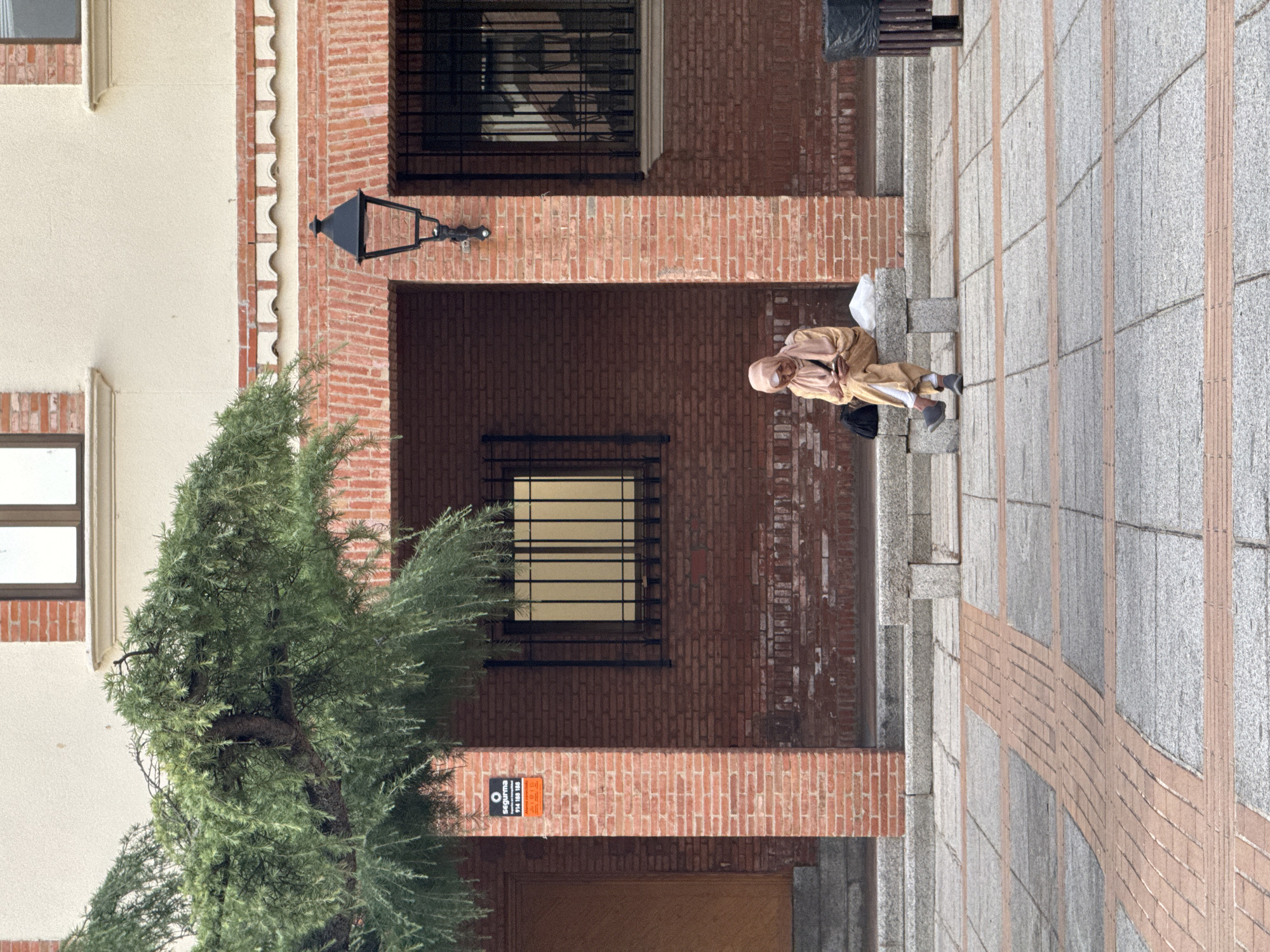 A woman wearing a light-colored headscarf and a matching outfit is sitting on a stone bench outside a brick building. She is looking down at her phone. Beside her on the bench are a black bag and a white bag. To her right, a black trash can is visible. The ground in front of the bench is paved with large gray tiles and is accented with bands of smaller orange bricks. The building behind her is made of red brick with several barred windows. A street lamp is mounted on the brick wall to the right of the woman. A large tree with green foliage hangs over the scene from the left. A security sign is affixed to a brick pillar on the left side of the image. The overall impression is an urban street scene in Las Rozas de Madrid, Spain.