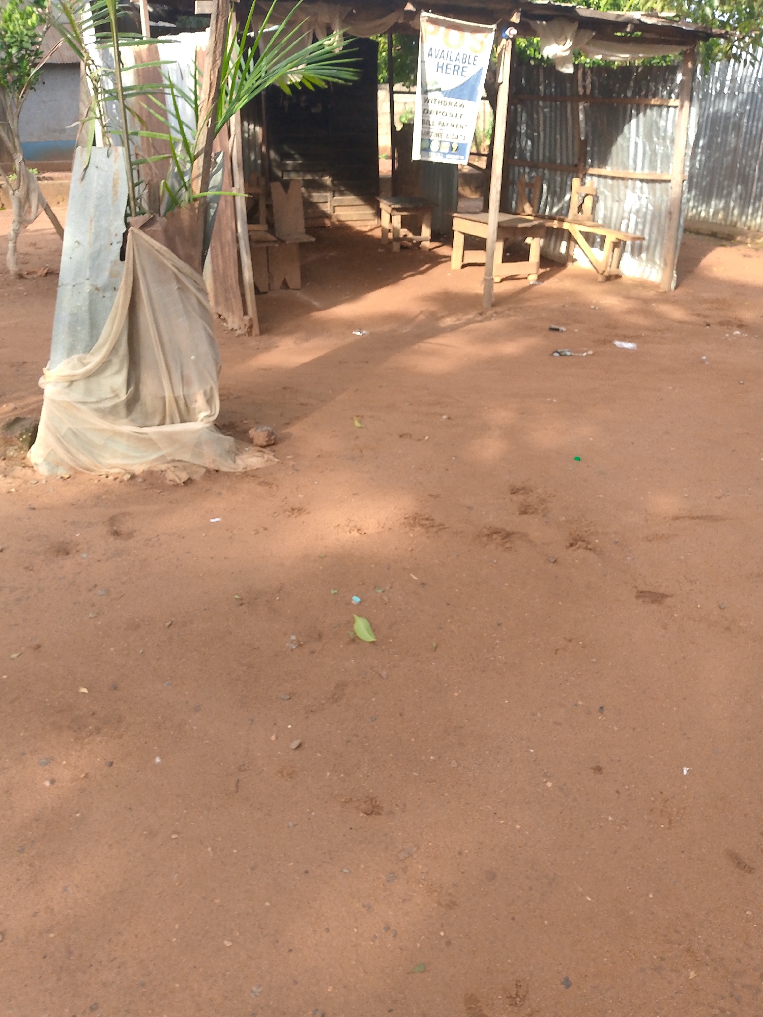 A wooden kiosk with a corrugated iron roof and walls stands on a dirt ground. A sign on the kiosk reads "AVAILABLE HERE WITHDRAWAL DEPOSIT BILL PAYMENT". Two wooden chairs and a table are placed inside the kiosk. A large plant with green leaves grows in front of the kiosk, partially covered by a piece of beige cloth. The ground is red soil and appears dry. In the background, there are other structures made of wood and corrugated iron, as well as trees. The overall scene suggests a small business or service point in a rural or semi-urban setting.