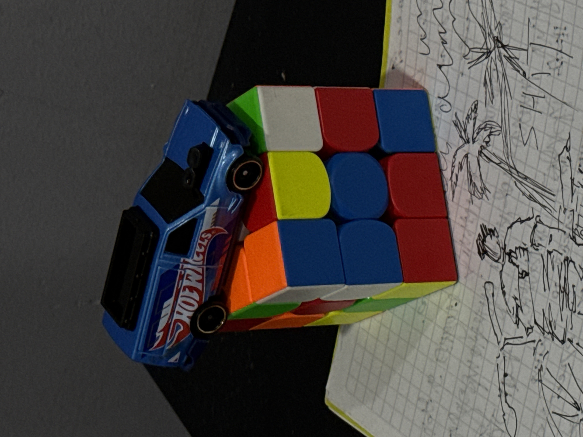 This image captures a still life arrangement featuring a toy car, a Rubik's Cube, and a piece of paper adorned with drawings. The objects are placed on a dark, possibly black, surface, with the gridded paper situated to the right.
A blue Hot Wheels toy car rests on top of a multi-colored Rubik's Cube. The car is positioned at an angle, with its front wheels elevated, leaning against the cube. The side of the car clearly displays the "HOT WHEELS" brand logo in white and red graphics. The Rubik's Cube itself shows several of its vibrant faces, including white, red, blue, yellow, green, and orange, indicating it is in a scrambled state.
Underneath the Rubik's Cube and extending to the right is a sheet of notebook paper with squared grids. This paper is covered with various hand-drawn sketches in black ink. Notable among these drawings are several palm trees, suggestive of a tropical climate. Next to one of the palm trees, partial text spelling "T H I S" can be discerned. Another significant drawing depicts a stylized human figure, possibly a man, with a beard and what appears to be a traditional Vietnamese conical hat (nón lá). The figure's posture suggests movement or carrying something.
The scene conveys no specific activity or event beyond the static display of these items. The lighting appears to be artificial and internal. While the image itself lacks explicit geographical markers, the presence of palm trees and the distinct conical hat on the drawn figure strongly align with the provided location context of Đà Nẵng, Vietnam.