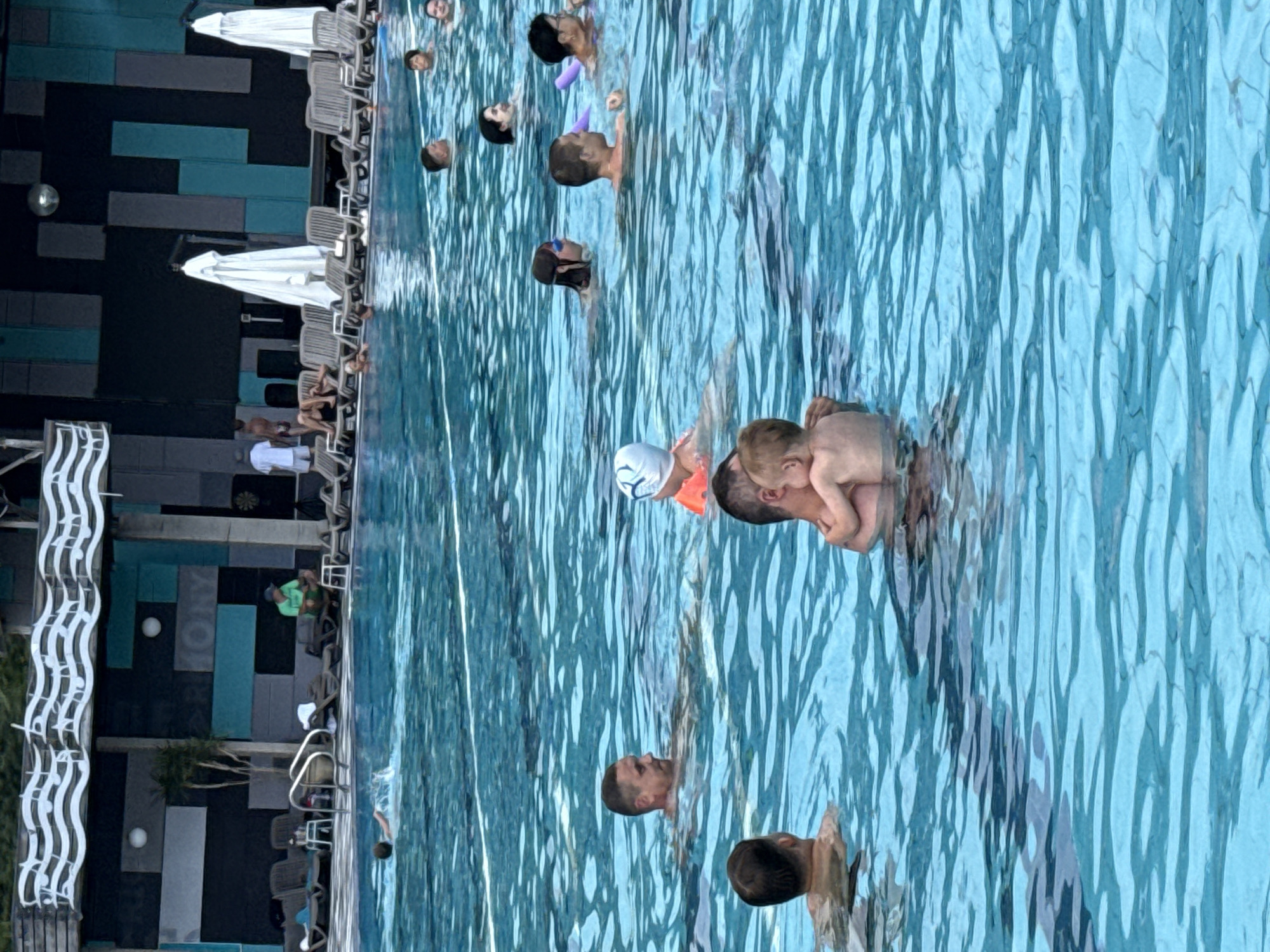 The image depicts a scene at an outdoor swimming pool, likely located in the Массандровский поселковый совет, Ukraine, given the context. The pool water is a bright blue, with visible ripples and some white lane markers. In the foreground, a man is holding a young child in his arms. The man is in the water, and the child appears to be resting against his chest. To the left of this pair, another man is swimming and looking to his right. Further back in the pool, several other individuals are visible, some swimming and others floating.
In the background, there is a structure with a dark wall decorated with rectangular panels of varying shades of blue and gray. Above this wall, there is a white decorative element that resembles musical notes. White lounge chairs are arranged around the pool, and large white umbrellas are visible. A few palm trees are also present near the pool's edge. A person in a green shirt is seated on a stool near the pool's edge, looking towards the pool. Several other people are seen sitting on lounge chairs in the background. The overall atmosphere suggests a recreational setting on a sunny day.