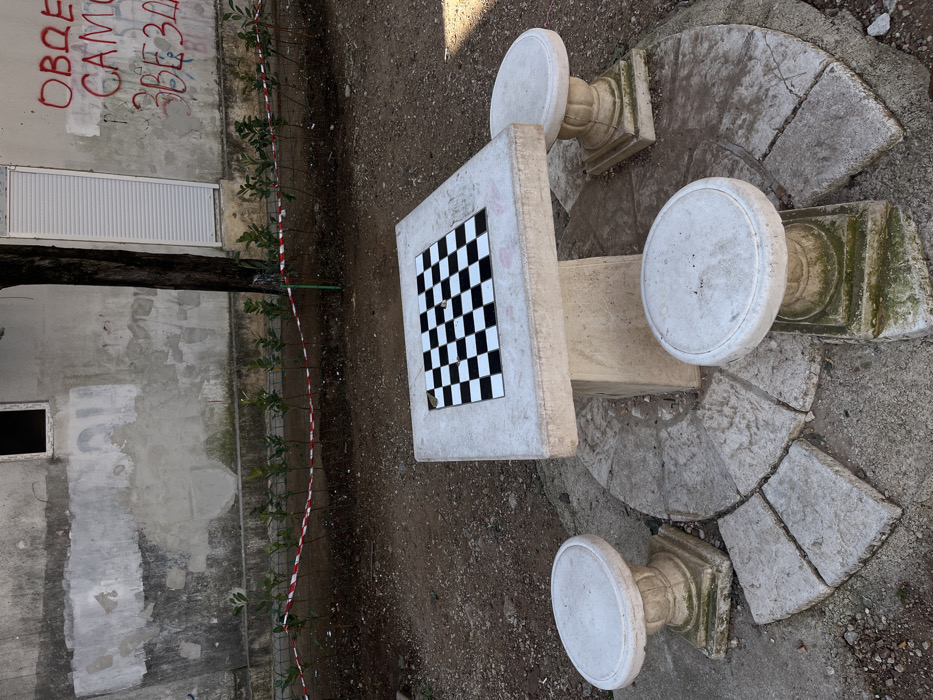 The image displays an unpopulated outdoor scene, located in the city of Budva, Montenegro. A stone chess table, featuring an embedded black and white tiled chessboard, is centrally positioned. Three matching weathered stone stools with fluted pedestals surround the table. These furniture pieces rest on a circular arrangement of broken, light-colored paving stones, while the immediate surrounding ground consists of dry, loose dirt and small gravel.
In the background, a heavily weathered, light-colored building wall exhibits extensive peeling render and visible repair patches. An open, dark rectangular window is visible on the upper left section of this wall. To its right, a dark tree trunk stands, partially obscuring a portion of the wall. Further right on the wall, a white vertical rolling shutter is installed. Red Cyrillic graffiti, including the words "ОВДЕ," "САМО," and "ЗВЕЗДА," is prominently spray-painted on the wall to the right of the shutter. A low barrier, formed by red and white striped caution tape strung between green posts, runs along the base of the wall, separating a row of small green plants from the foreground. No individuals or active interactions are present in the scene.