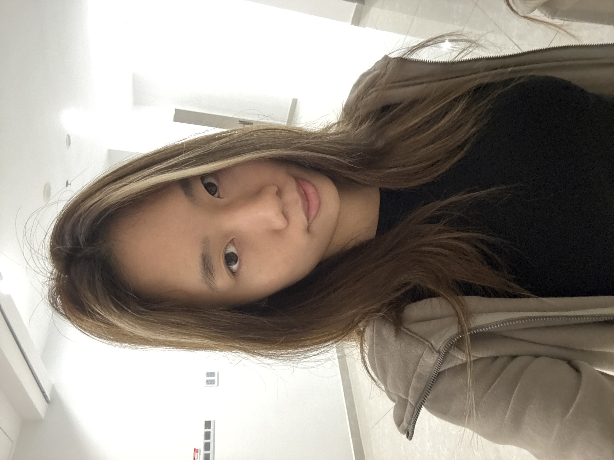 A young woman with long brown and blonde hair and wearing a black shirt and beige hoodie poses indoors. The background appears to be a brightly lit hallway with a white tile floor and walls. A panel with some red and white rectangles is visible on the wall. The location is implied to be Nan'an District, China.