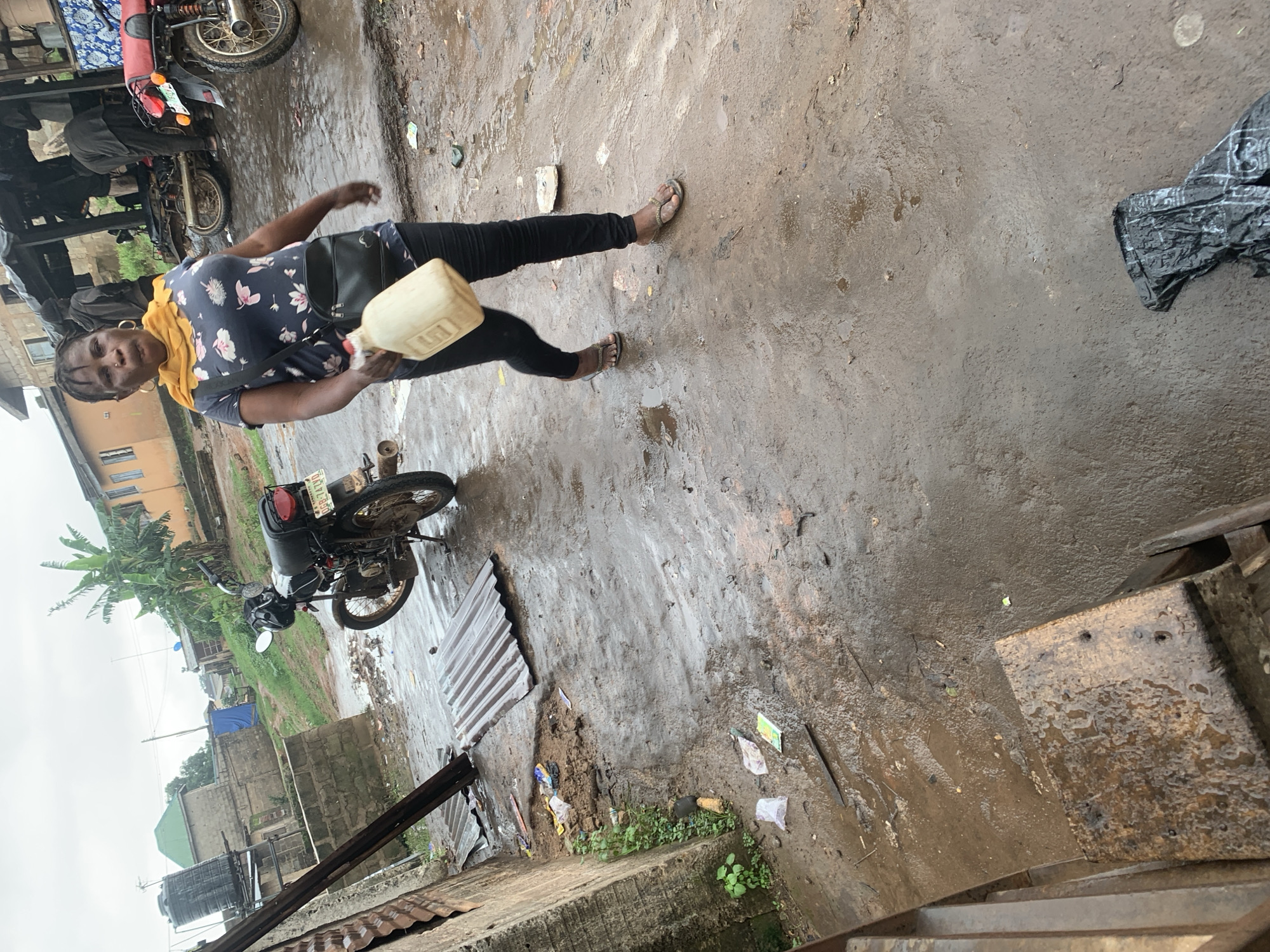 In Shagamu, Nigeria, a woman walks across a wet, muddy street, holding a beige plastic jug. She is wearing a dark blue t-shirt with a floral pattern and black leggings, and a yellow scarf around her neck. She has a black crossbody bag. The street is littered with debris, including plastic bags and corrugated metal sheets. To the left of the woman, a motorcycle is parked. In the background, there are buildings, a water tank, and more motorcycles. The sky is overcast.