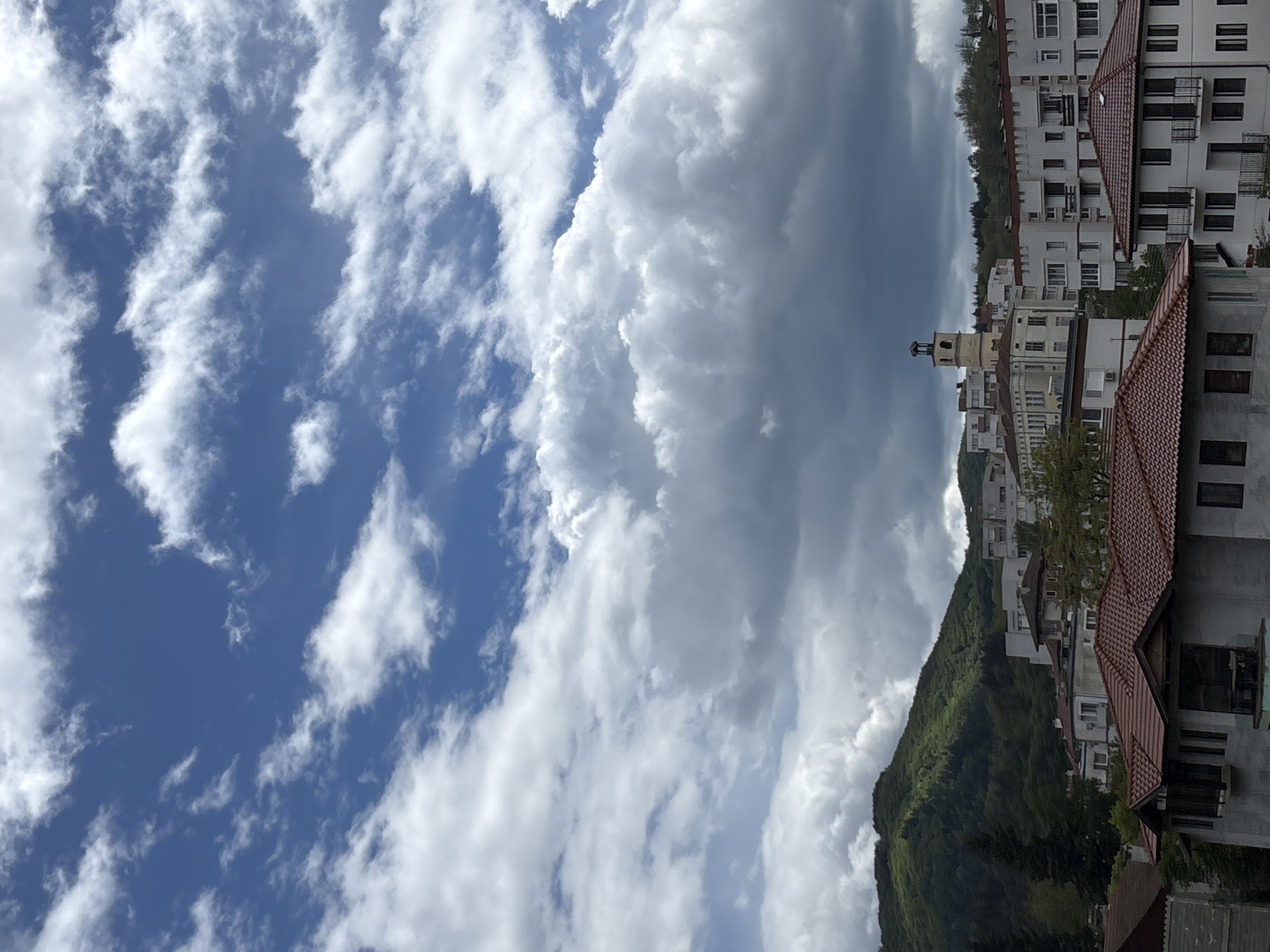 The image shows a view in Zlatograd, Bulgaria. The sky is predominantly blue with wispy cirrus clouds on the left side, transitioning to large, dense cumulus clouds in the center and right. These clouds indicate potentially unsettled weather.
On the right side of the image, buildings with white facades and red-tiled roofs are visible, ascending a hillside. A distinctive tower with a pointed roof is situated among the buildings. The hillside is covered with lush green vegetation. The photograph seems to be taken in a slightly elevated position.