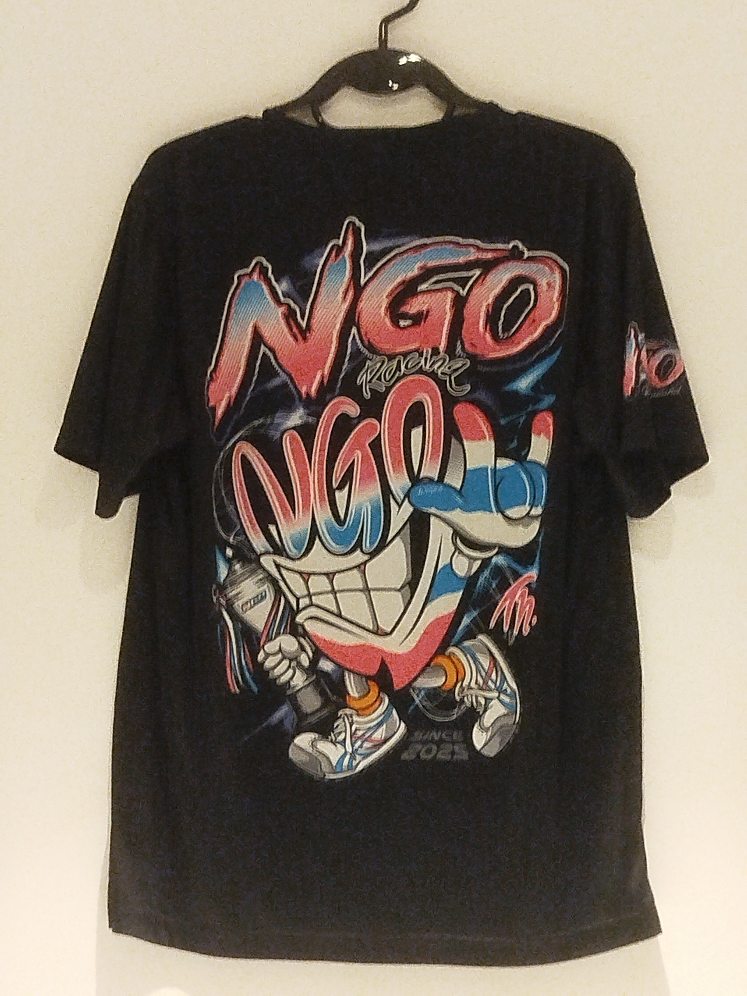 A black t-shirt with a graphic print is hanging on a black hanger against a white wall. The graphic on the t-shirt depicts a cartoon character with a large, smiling mouth and wide eyes. The character is wearing a white glove with blue stripes on its hand and is holding a can of spray paint. The t-shirt has the text "NGO RACING" written in large, colorful letters above the character. Below the character, the text "SINCE 2023" is visible. The overall aesthetic of the graphic is bold and vibrant, with a street art or graffiti style. The t-shirt is the central focus of the image. The location is not provided within the image or accompanying information.