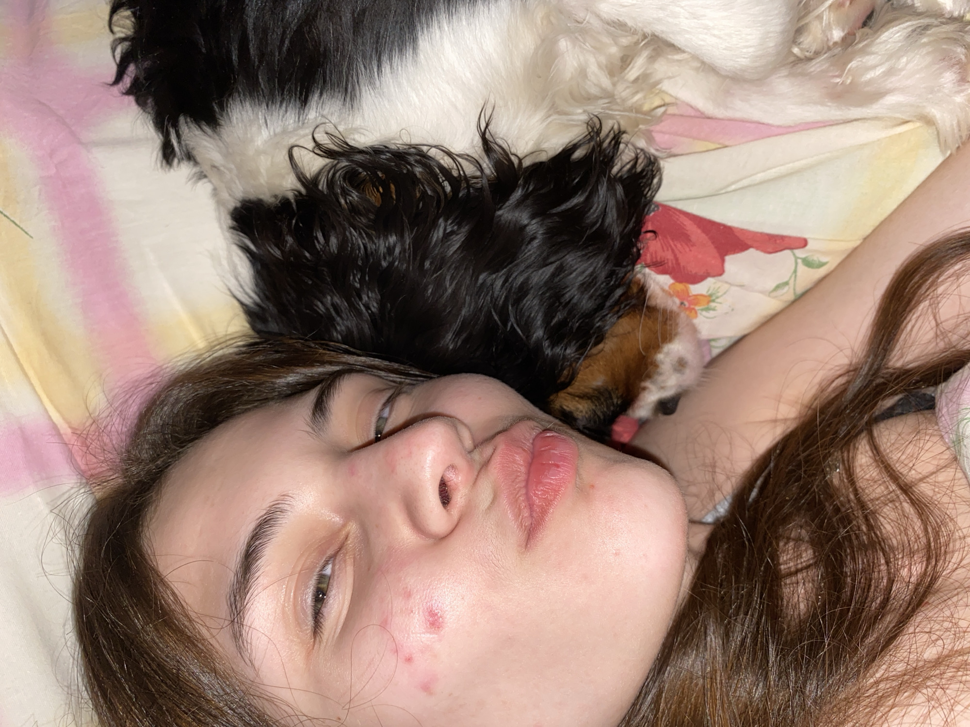 The image shows a close-up, overhead view of a young person and a dog resting together on a bed in Votkinsk, Russia.
The main subject is a young person, likely female, positioned in the foreground on their back, looking towards the camera. They have long brown hair that spreads out on the bed. Their lips are slightly pursed, and there are a few blemishes visible on their cheek. Next to and slightly behind the person's head, a dog with black, white, and brown fur is also lying down. The dog's body is mostly out of frame, but its head is visible with its nose and a hint of its tongue near the person's ear.
Both subjects are on a bed covered with a patterned sheet or blanket. The bedding features a combination of soft pink and yellow stripes, along with a floral motif displaying red flowers and green leaves. The scene captures a peaceful and intimate moment of companionship between the person and their pet, suggesting relaxation or lounging indoors. The time of day is not discernible from the lighting, and there is no visible text in the image.
