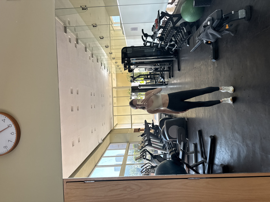 This image depicts the interior of a modern fitness center or gym located in Mexico City, Mexico. The scene is dominated by a large wall-mounted mirror, which reflects a lone female subject taking a selfie with a smartphone held in her right hand, obscuring her face. She is dressed in a light-colored crop top or sports bra, black athletic leggings, and light-colored athletic shoes.
The gym features dark, likely rubberized, flooring, light beige walls, and a light-colored ceiling with square recessed lighting fixtures. A round wooden-framed wall clock, displaying approximately 4:45, is mounted above the mirror. A prominent wall of large windows extends across one side of the facility, providing natural light and an exterior view of an urban landscape, which includes several modern tall buildings and green trees characteristic of Mexico City.
Various exercise equipment is present throughout the gym. Cardio machines such as treadmills, elliptical trainers, and stationary bikes are positioned near the windows. Strength training equipment includes a multi-station cable machine, a two-tiered rack stocked with multiple sets of dumbbells, and an adjustable weight bench with a dark grey towel or garment draped over it. Additional fitness accessories observed are a large blue stability ball near the cardio area, and a large green stability ball and a black foam roller situated near the dumbbell rack. No other individuals are visible or actively using the equipment.