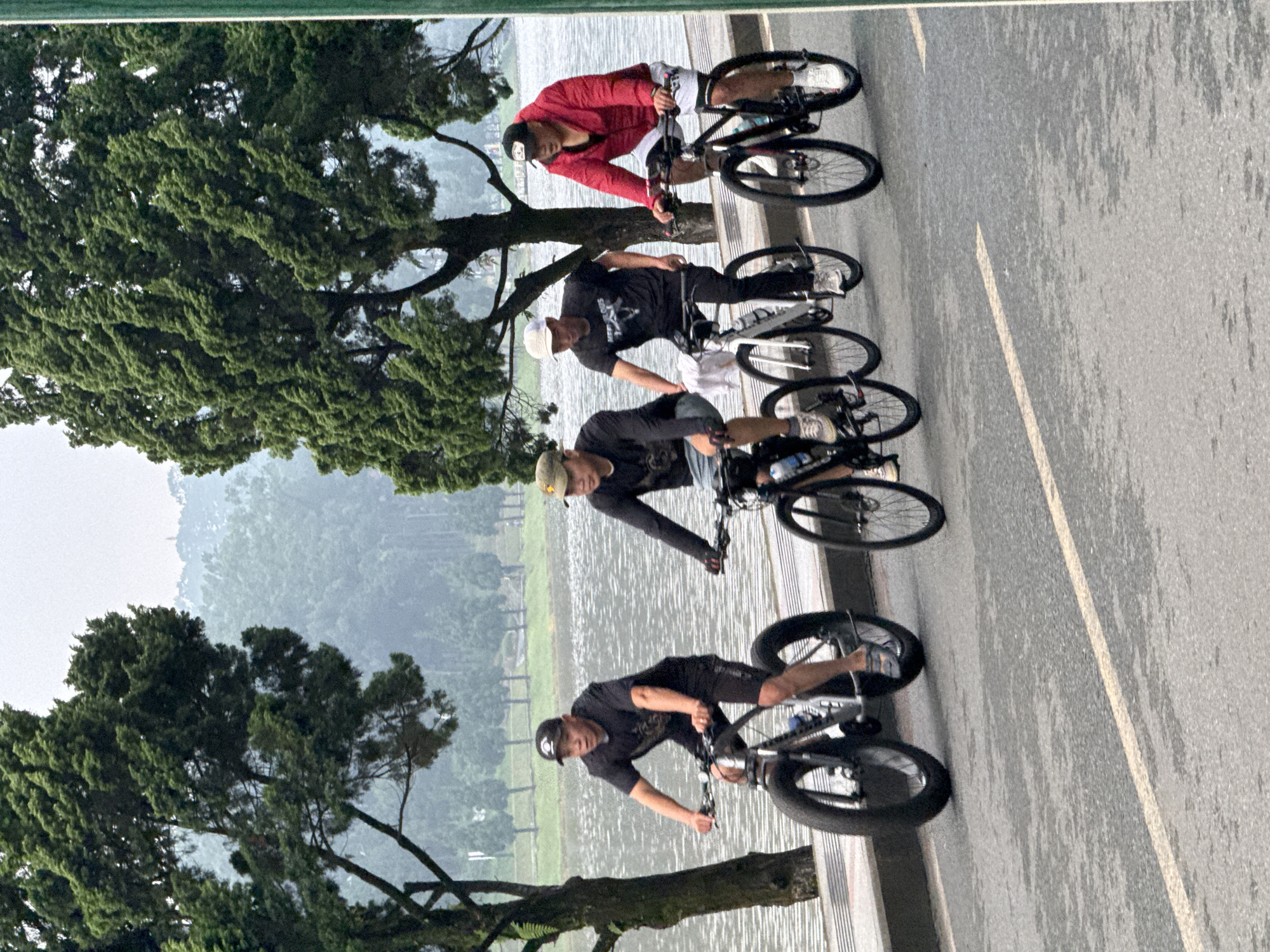 Four individuals are cycling along a paved path next to a body of water, likely Xuan Huong Lake in Da Lat, Vietnam, as indicated by the surrounding cypress trees and misty mountainous backdrop. The first cyclist from the left wears a black t-shirt, black shorts, and a black baseball cap, riding a fat-tire bicycle. The second cyclist, to his right, is dressed in a black t-shirt, black pants, and a tan baseball cap, also on a bicycle. The third cyclist is wearing a black t-shirt and white cap, and the fourth, on the far right, sports a red jacket, white shorts, and a black baseball cap, riding a dark-colored bicycle. The pavement shows a light yellow line, suggesting a marked lane. The overall atmosphere is overcast, and the water appears calm.