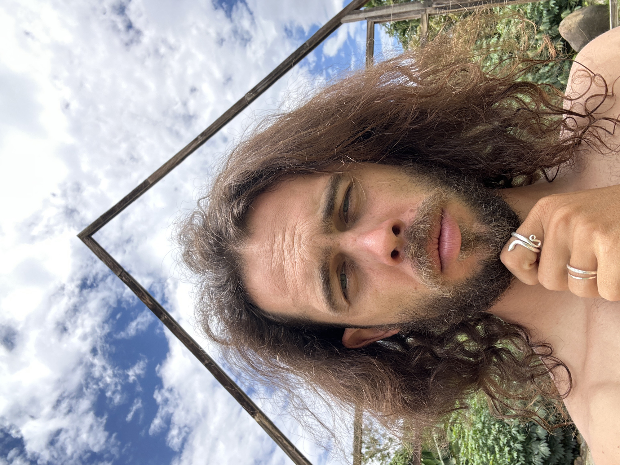 A frontal, selfie-style portrait of an adult male is captured outdoors in Nghia Lo Ward, Vietnam. The subject has long, dark brown, wavy hair that falls past his shoulders, a full beard, and a mustache. His eyes are light-colored, appearing greenish-hazel, and he maintains a direct gaze at the camera with a neutral to slightly contemplative expression. Subtle horizontal lines are visible on his forehead. His right hand is raised, with fingers curled and the thumb resting beneath his chin, revealing two silver rings: a plain band on his ring finger and a coiled, serpentine-like ring on his index finger. His upper torso and shoulders are bare. In the background, an open, weathered wooden A-frame structure is prominently positioned, framing the sky above and around his head. The sky is bright blue with scattered white cumulus clouds. Green foliage and distant trees are visible in the lower background, particularly to the right and left of the subject. The scene is illuminated by natural daylight.