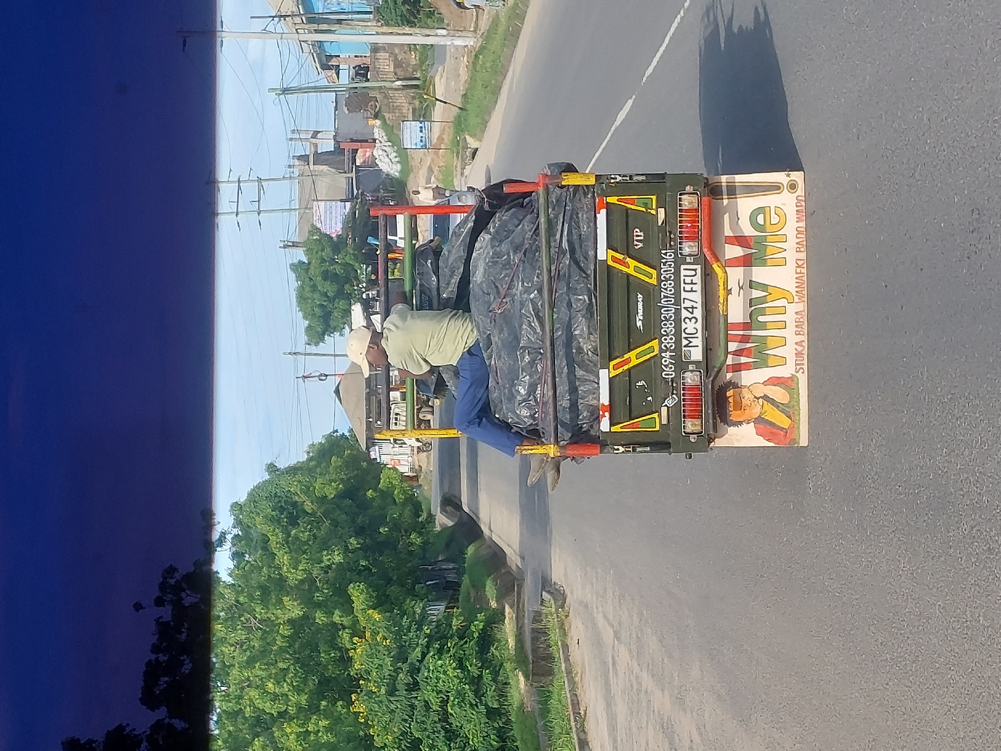 The image shows a modified truck driving on a paved road, likely within Dar es-Salaam, Tanzania, given the phone numbers on the vehicle. A man is precariously riding on the back of the truck, exposed. The truck bed is covered with a black tarp over what appears to be various items. The rear of the truck has signage that reads "Why Me!" and includes other smaller text. The surrounding environment includes trees, buildings, and power lines. The image is taken from another moving vehicle, as suggested by the blue tint in the left foreground.