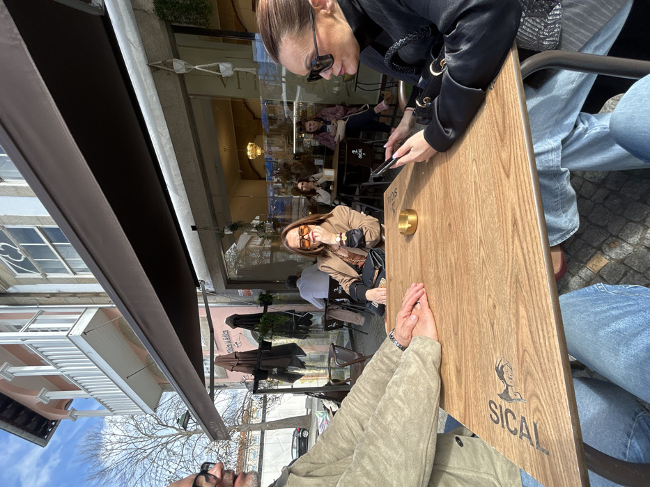 The image depicts an outdoor cafe scene on a sunny day in Chaves, Portugal. Three individuals are seated around a light-colored wooden table with a "SICAL" brand logo and a stylized female figure. A small, golden cylindrical ashtray rests on the table.
On the left, partially visible, is a male wearing glasses, a light-colored jacket, and blue jeans, with his hands clasped on the table. In the center, a female wearing brown sunglasses, a tan blazer over a patterned top, and a watch on her left wrist is looking directly towards the camera, with her right hand near her chin. A black shoulder bag is positioned next to her. On the right, a female in profile, wearing dark sunglasses and a black top, is engrossed in a smartphone held with both hands; a black bag with a braided handle is visible.
Behind the primary subjects, an outdoor seating area is covered by a brown awning. Additional dark-colored cafe tables and chairs are visible, occupied by other patrons. Through a large glass storefront, the cafe's interior reveals more customers and a warm light fixture. Hanging plants in macrame holders adorn the storefront. A sign "MÓVEL - ÓTICA" is visible under the awning.
The background shows a street with a bare deciduous tree and a parked black sedan. Historic buildings line the street, including one with a pink facade and a white balcony supported by columns, and an adjacent building featuring a light-colored stone or stucco facade with ornate windows, including a distinct quatrefoil design. The sky is clear and blue with scattered clouds, indicating bright daylight and strong shadows.