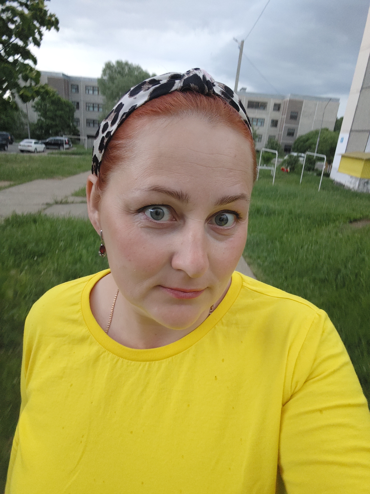 A woman is taking a selfie outdoors. She has red hair, a leopard print headband, and a yellow t-shirt. She is wearing earrings and a necklace. The background shows a grassy area with buildings, trees, and playground equipment. It is daytime and the sky is overcast. The woman is located in the city of Марковский, Russia.