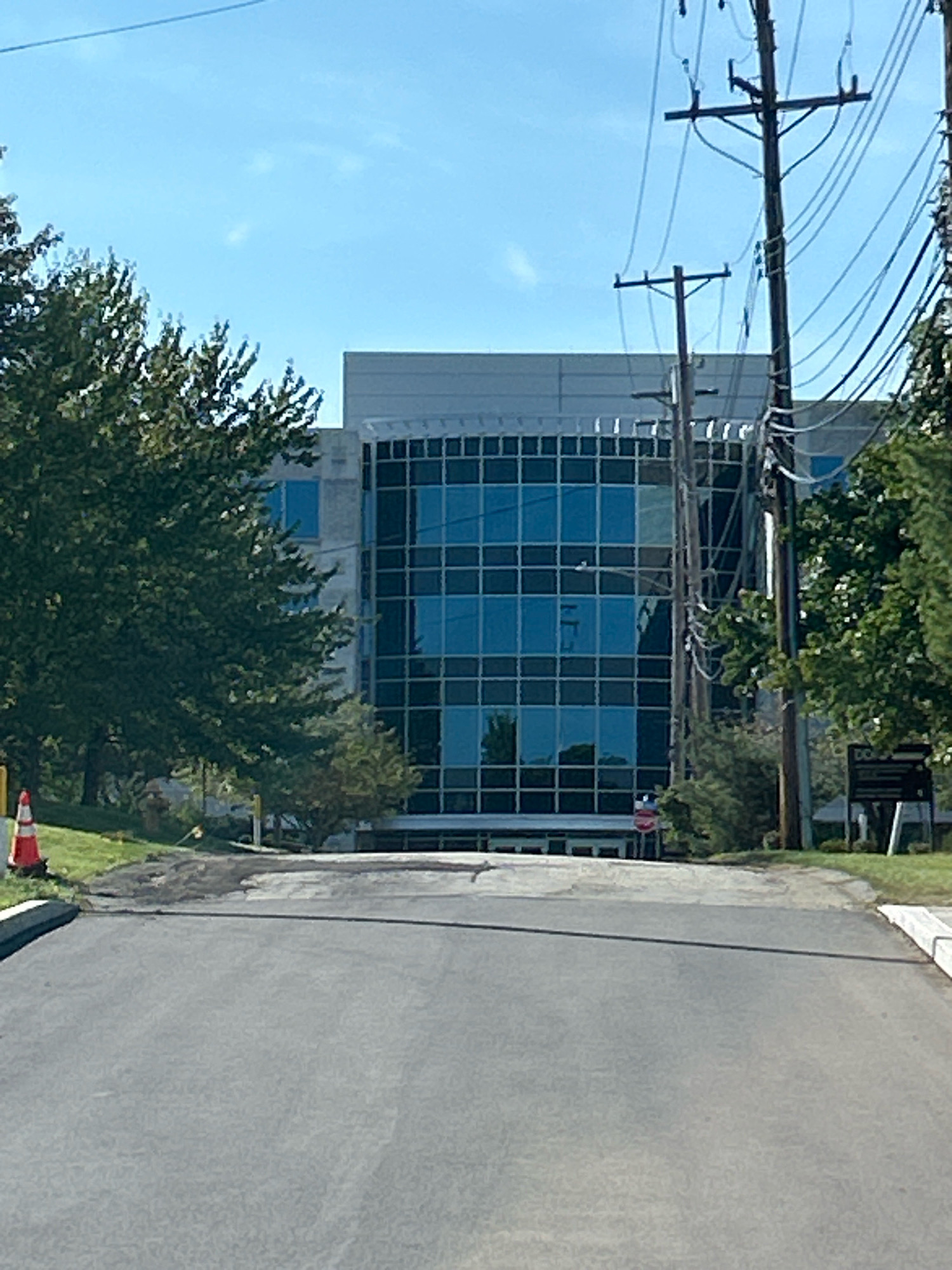 The image depicts a large, modern office building with a glass facade, situated in Collier Township, United States of America. The building is bordered by trees and utility poles with a network of wires. In the foreground, a paved road leads towards the building, with a traffic cone visible on the left side of the road. The sky is clear and blue.