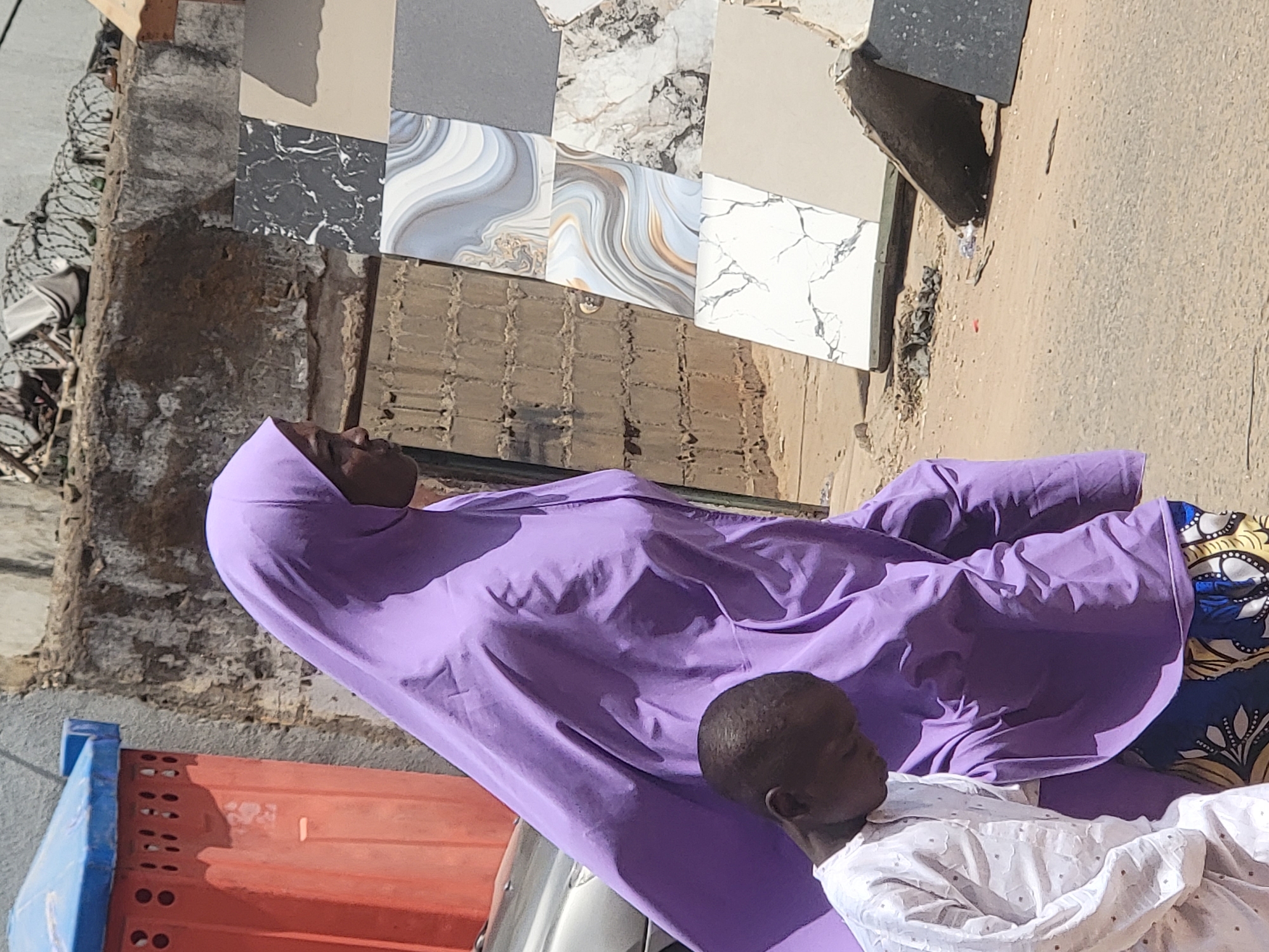 A person wearing a purple hijab is lying down outdoors, possibly on a cot. A child, dressed in white, is next to them. They are on a dirt surface. Nearby are several tiles of various colors and designs leaning against a wall. An orange and blue object is partially visible in the lower left corner. The location appears to be an urban setting. The location is in the city of Maiburiji, Nigeria.