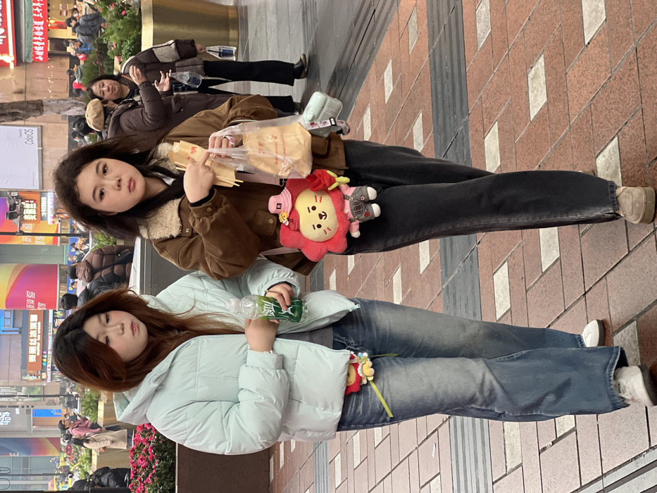 Two young women stand on a brick-paved pedestrian street. The woman on top wears a brown jacket with a light-colored lining, black pants, and beige shoes. She holds a plastic bag with what appear to be food skewers. The woman below wears a light blue puffer jacket, blue jeans, and white shoes, holding a plastic bottle. Both women have plush toys attached to their clothing. In the background, there are signs, buildings, and people walking by.