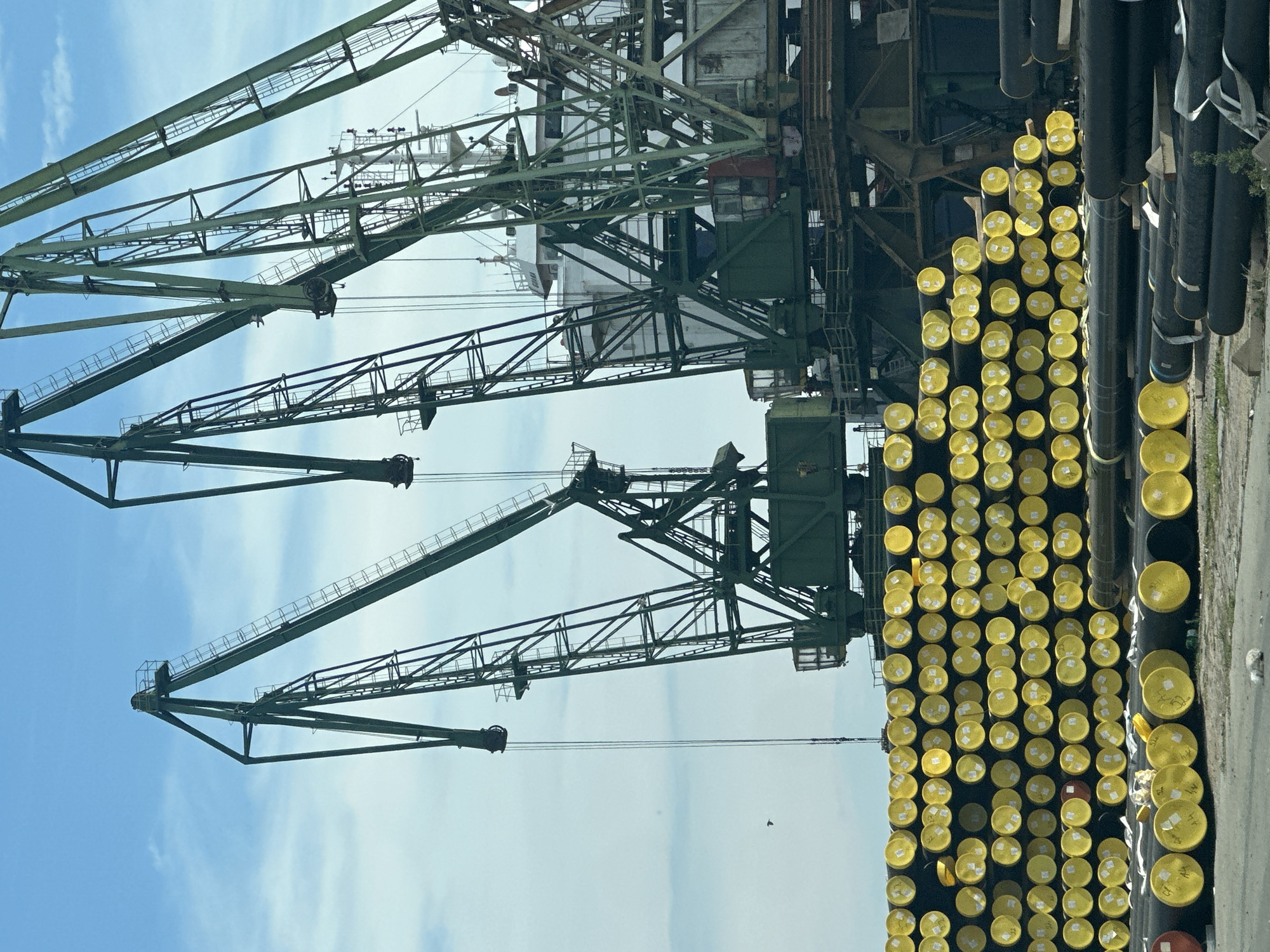 In an industrial port, large green gantry cranes dominate the skyline against a backdrop of a partly cloudy blue sky. In the foreground, stacks of large black pipes with bright yellow caps are neatly arranged. The cranes appear to be in a state of disuse or preparation for work. A white ship is visible behind the cranes, partially obscured. The scene suggests a location where heavy industrial materials are stored and handled for shipment.