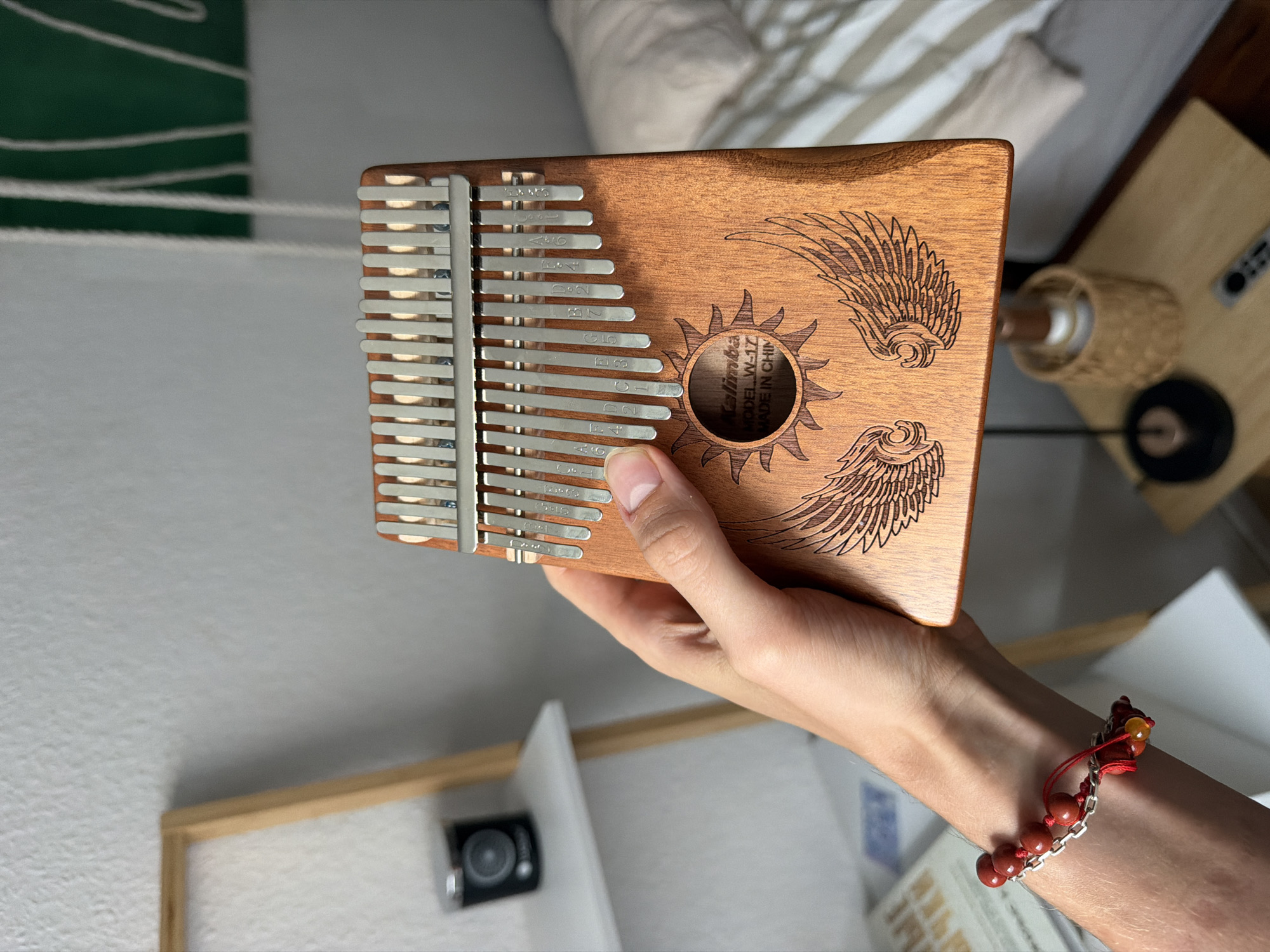 A hand holds a wooden kalimba, a thumb piano, in front of a blurred background. The kalimba has metal tines and is engraved with a sun and wings design. The background shows a shelf with a black candle, a bed with striped bedding, and a nightstand with a lamp and remote control. The scene appears to be indoors, in a bedroom. No specific location in Herne, Germany is identifiable.