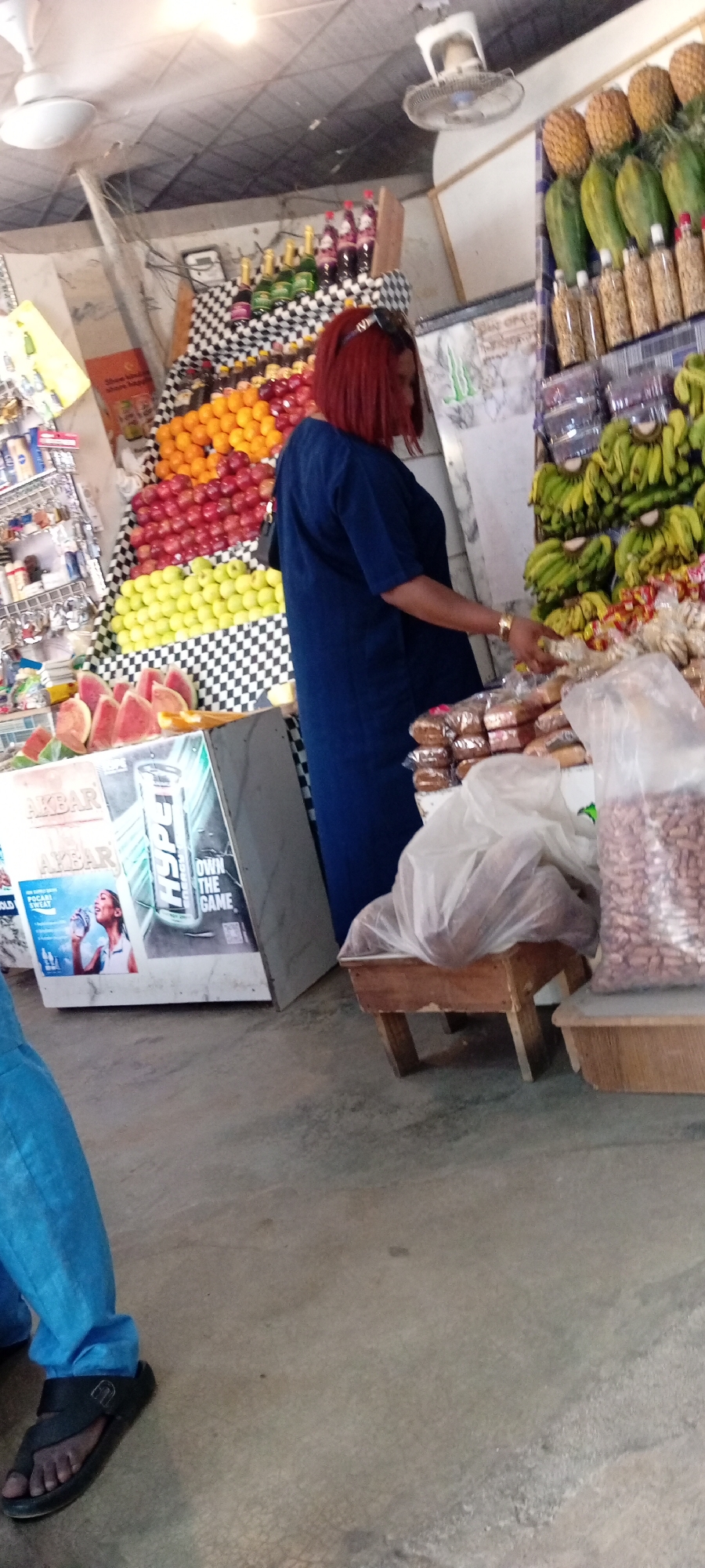 The media file depicts a vibrant indoor market scene, likely in Aviation Quarters, Nigeria. A woman with striking red hair is the central human subject, wearing a long, dark blue dress and a gold bracelet. She is actively engaged, reaching out towards a display of packaged goods. Her posture suggests she is either examining or selecting items.
The setting is a market stall overflowing with a diverse array of products. Fruits are prominently displayed, including a colorful arrangement of oranges, red apples, and yellow apples, stacked against a black and white checkered backdrop. Sliced watermelons are visible at the front left of the stall. Above and to the right of the woman, bunches of green bananas are neatly arranged, and a display of pineapples is positioned at the top. Various bottles, likely beverages or oils, are also present. The stall also features packaged goods, including what appear to be snacks or bread, some in clear plastic bags, and a large bag of what seems to be nuts or beans.
To the left of the woman, a white display box showcases advertisements. One prominent advertisement features the brand "AKBAR" twice and an image of a woman drinking from a bottle with the text "POCARI SWEAT". Another advertisement on the same box prominently displays "HYPER" and "OWN THE GAME".
The floor is concrete, and the overhead lighting is bright, suggesting it's daytime. The overall impression is one of a bustling market, with a focus on fresh produce and packaged goods. The woman's engaged stance implies a transaction or shopping activity. The scene is lively and visually rich due to the variety of colors and textures.