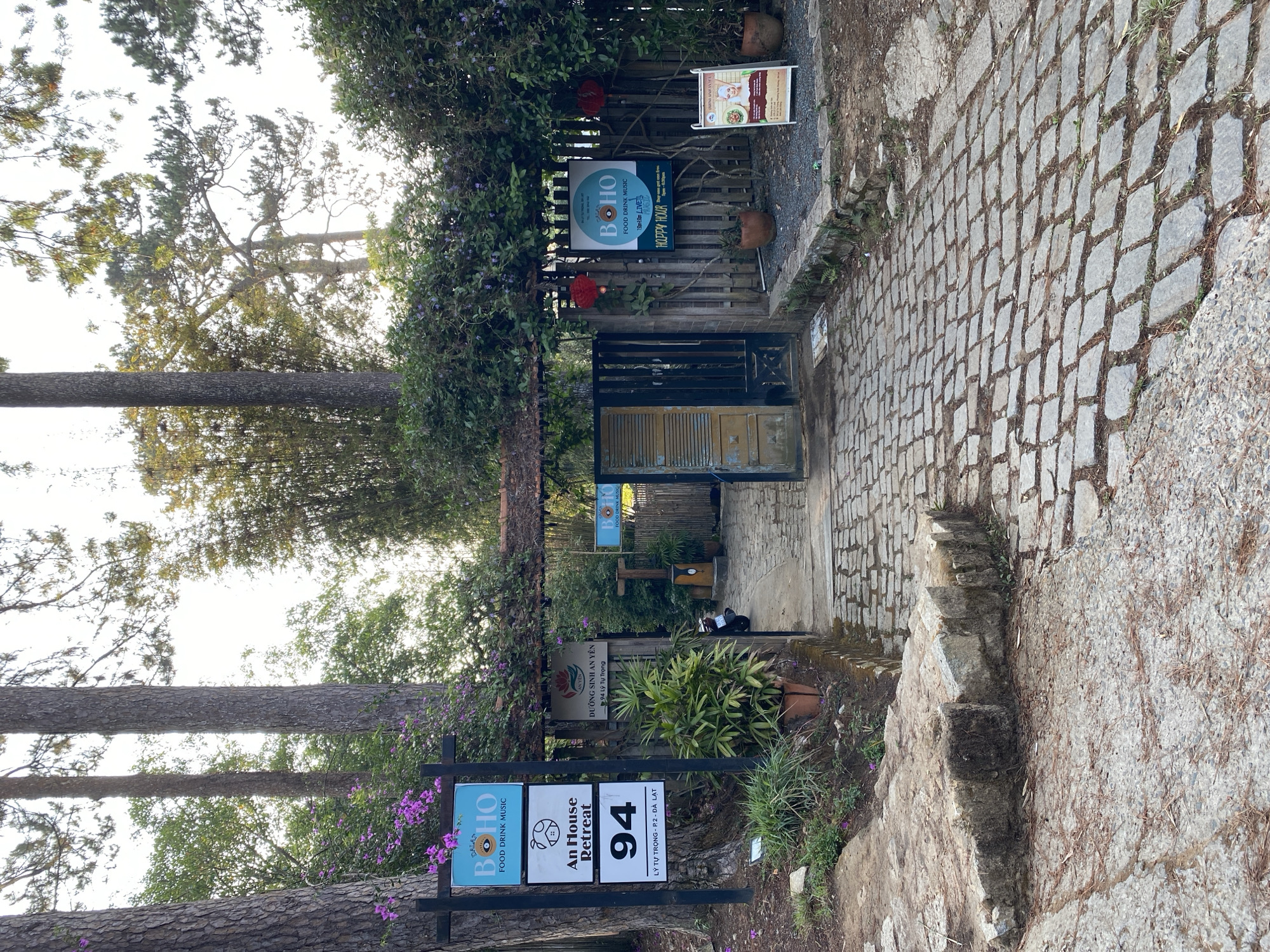 The image shows an entrance to "An House Retreat" located at 94 Ly Tu Trong Street in Da Lat, Vietnam. A stone path leads through a gated entrance covered in foliage. Signs indicate "Boho Food Drink Music Live" and "Happy Hour." A sign also denotes "Dưỡng Sinh An Yên", with the address. The surrounding area includes trees and additional foliage. The entrance features a wooden gate with an opening. The foreground shows stone steps and pavers leading up to the entrance.
