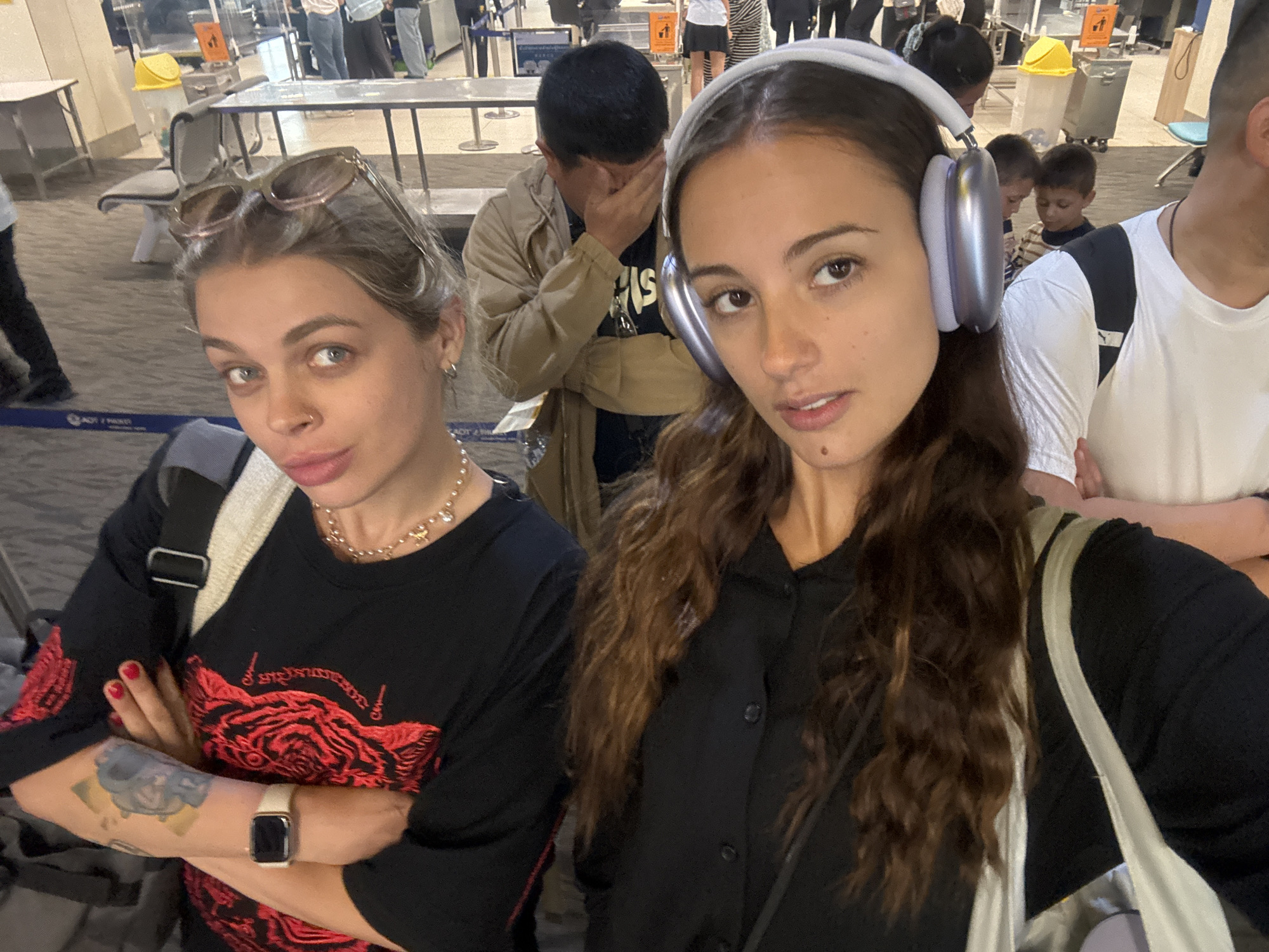 The image depicts two young women taking a selfie indoors, likely in an airport terminal or security area. The woman on the left has light brown hair, sunglasses perched on her head, and a nose piercing. She sports a slightly playful expression and wears a black long-sleeved shirt with an intricate red design, possibly a stylized animal or script. A gold-colored necklace adorns her neck, and a white smartwatch is visible on her left wrist, above a tattoo on her forearm. Her arms are crossed.
Beside her, the woman on the right has long, dark, wavy hair and wears silver over-ear headphones. She looks towards the camera with a neutral gaze, dressed in a dark button-up shirt, and carries a light-colored shoulder bag.
In the blurred background, several other people are visible. A man directly behind the two women covers his face with his hand. Further back, two small children can be seen. The setting features grey patterned flooring, metal barriers, and tables, consistent with an airport environment. A yellow bin is present in the mid-ground. Partially visible on a blue sign on the floor are the words "AOT" and "MAI KHAO," confirming the location as Tambon Mai Khao, Thailand (home to Phuket International Airport). The scene suggests a candid moment during travel.