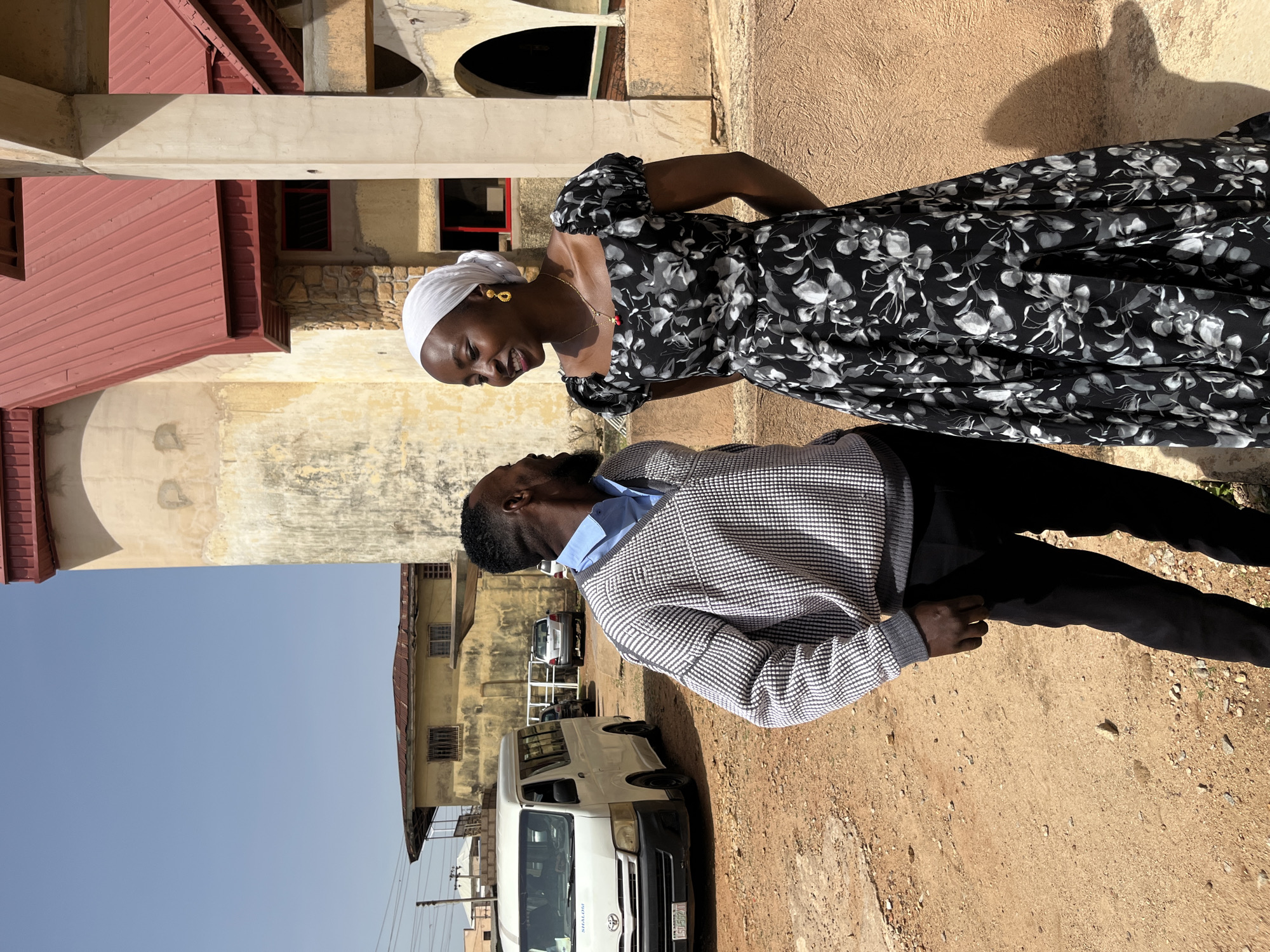 A man and a woman are standing outside on a dirt road in Bukuru, Nigeria. The man, wearing a checkered sweater over a blue shirt and black pants, is looking at the woman. The woman, in a black floral dress and white headscarf, is smiling at the man. Behind them, a white van is parked, and in the background, there are buildings with red roofs and a light-colored, textured wall. The sky is clear and blue.