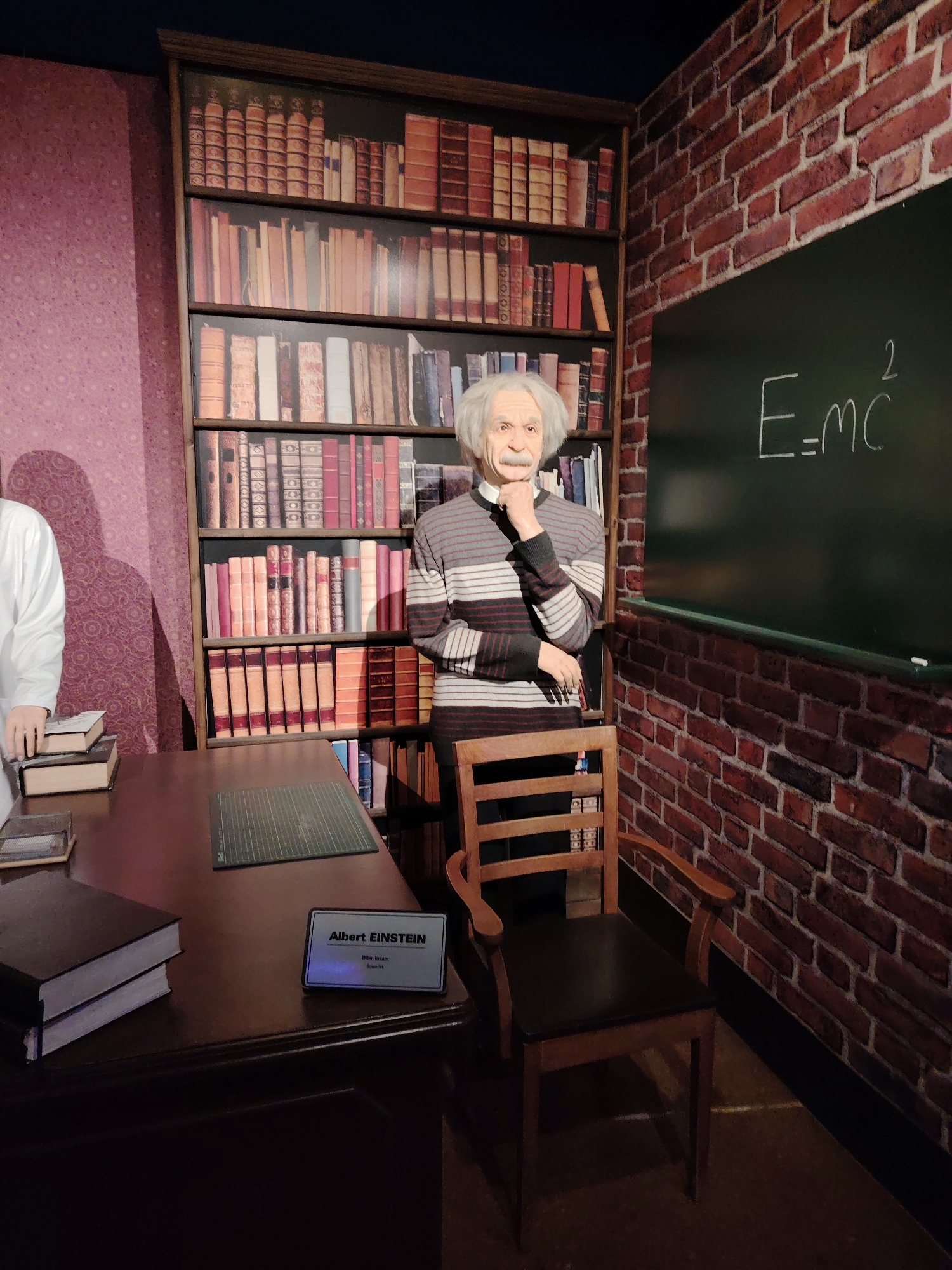 This image captures a wax figure exhibit, likely from a museum in Eskisehir, Turkey, depicting a scene reminiscent of Albert Einstein's study or office.
The main subject is a life-sized wax figure of Albert Einstein, positioned centrally. He is portrayed with his characteristic wild white hair and mustache, wearing a horizontally striped sweater in black, white, and red tones. His right hand is raised to his chin in a contemplative pose.
Behind the Einstein figure stands a tall, dark wooden bookshelf packed with numerous old-fashioned books. To his right, a section of wall is covered in red brick, against which a dark green blackboard is mounted. The iconic equation "E=MC²" is prominently written on the blackboard in white chalk.
In the foreground, a dark wooden desk extends across the lower left part of the frame. On the desk, there are two stacks of books, and a green cutting mat. A silver nameplate is placed on the desk, clearly displaying the text "Albert EINSTEIN" at the top, followed by "Bilim Insanı" (Turkish for Scientist), and "Scientist" below it. A wooden chair with a dark seat and armrests is positioned next to the desk, in front of the brick wall. Part of another figure, wearing a white coat, is visible on the far left, standing beside the desk.
The wall on the left side of the scene features a patterned purple wallpaper. The lighting is consistent with an indoor museum setting, creating a detailed and immersive display of the famous physicist.