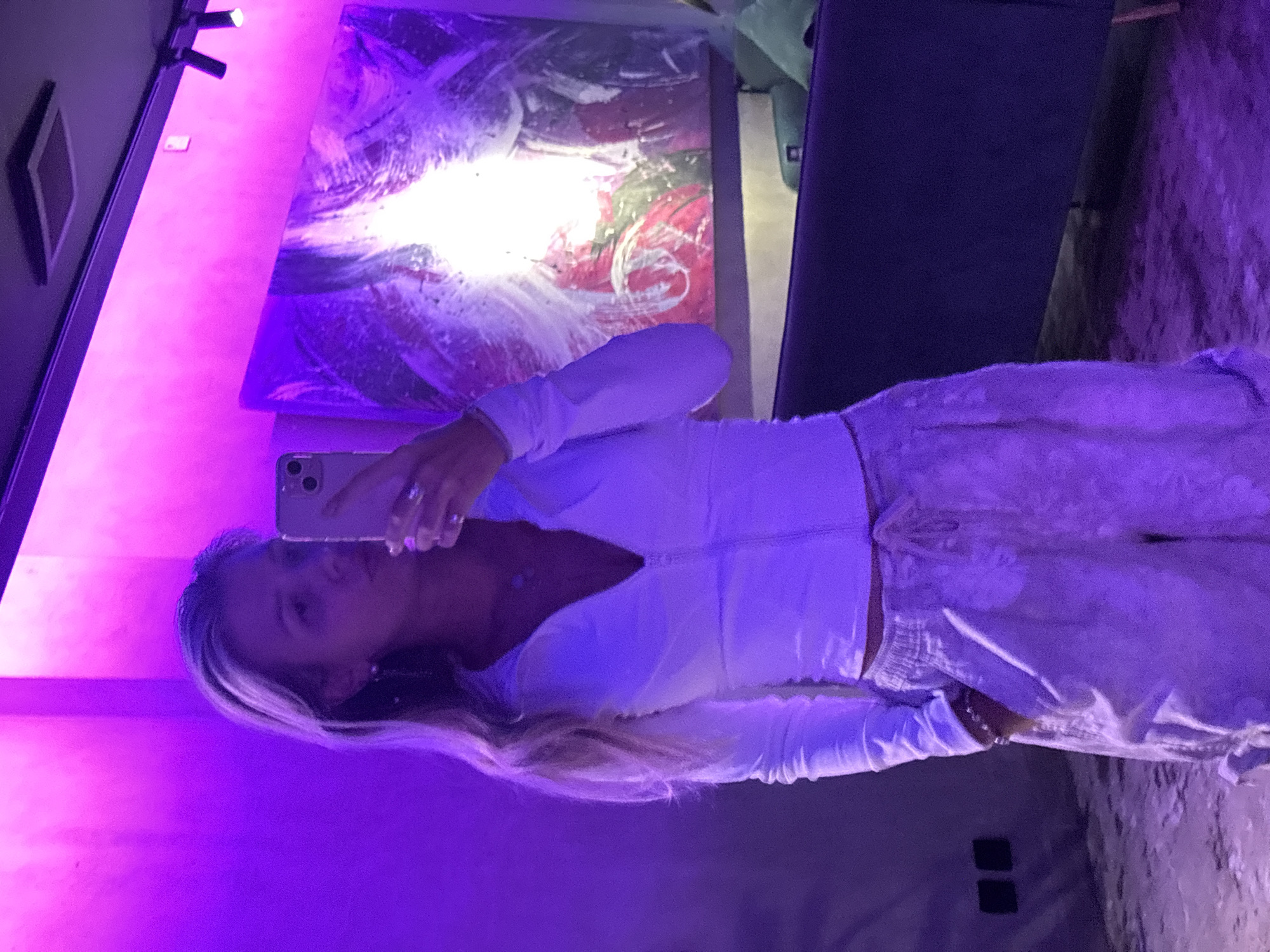 A young woman, appearing to be in her early twenties, stands in an indoor environment illuminated by purple and pink lighting. She is taking a selfie in a mirror, holding up a smartphone. She is wearing a white zip-up top and loose-fitting patterned pants. Her hair is long and blonde, styled in waves. A large, abstract painting hangs on the wall behind her, and a dark couch is visible in the background. The floor appears to be a textured, light-colored surface. Two dark electrical outlets are visible on the left wall.