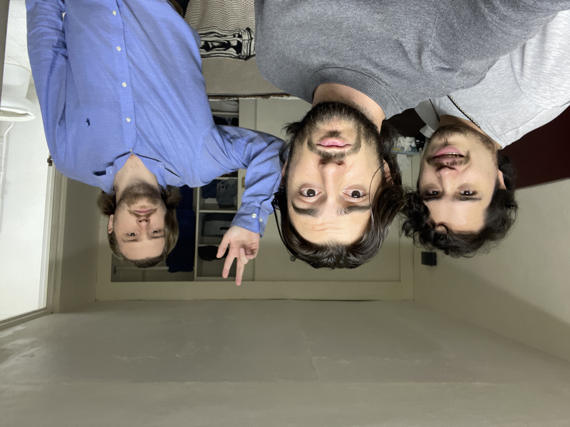 Three men are positioned upside down, likely taking a selfie. The man on the left is wearing a blue button-down shirt. The man in the middle is wearing a gray sweater and has a headset. The man on the right is wearing a light gray shirt. They appear to be in a room with white walls and a closet in the background. The location is unknown and there is no way to tell this is in Huixquilucan de Degollado, Mexico.